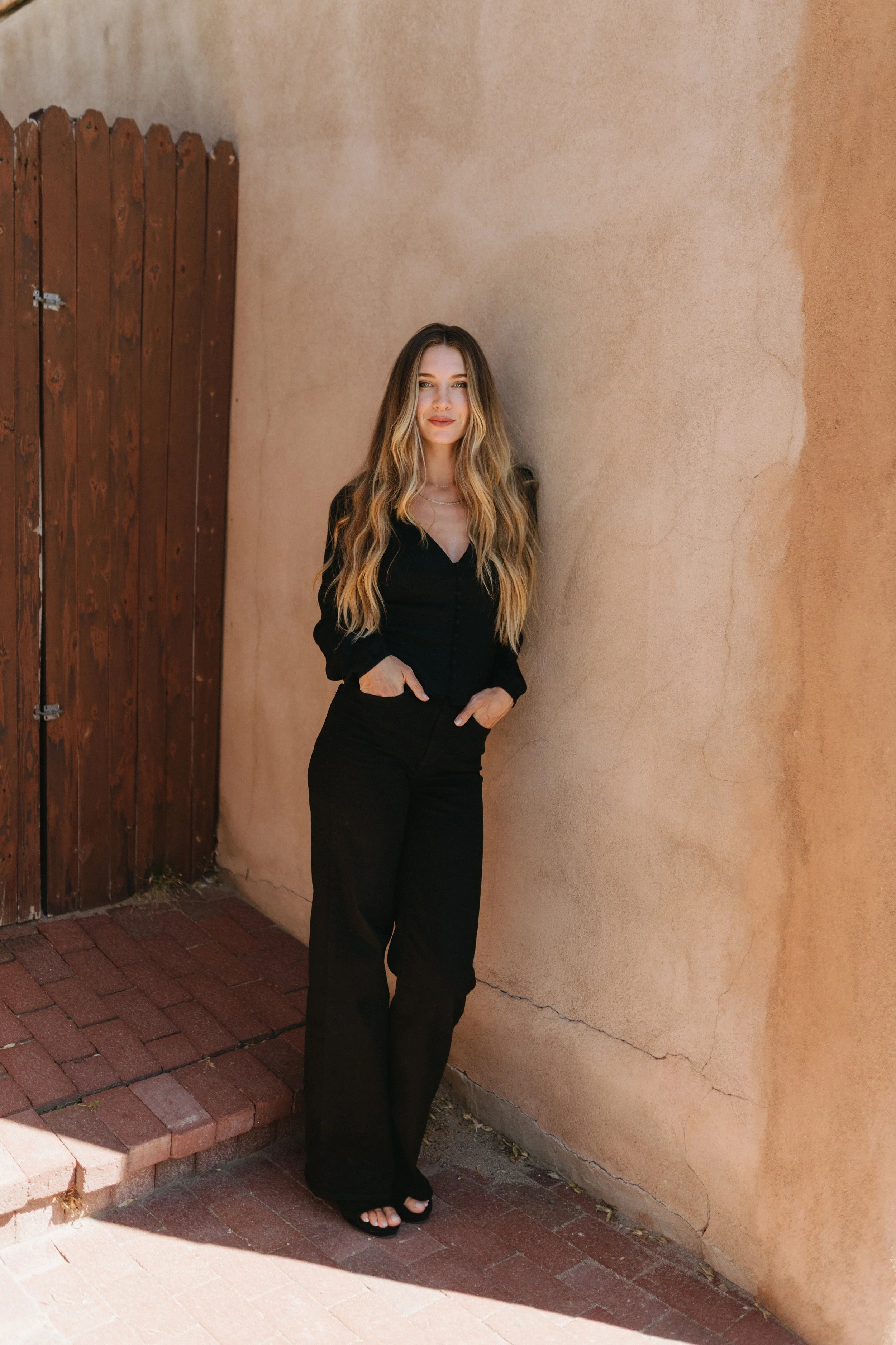 A woman, Nadia Mousleh, in New Mexico with long wavy blonde hair in black clothing standing outdoors against a beige wall and wooden gate.