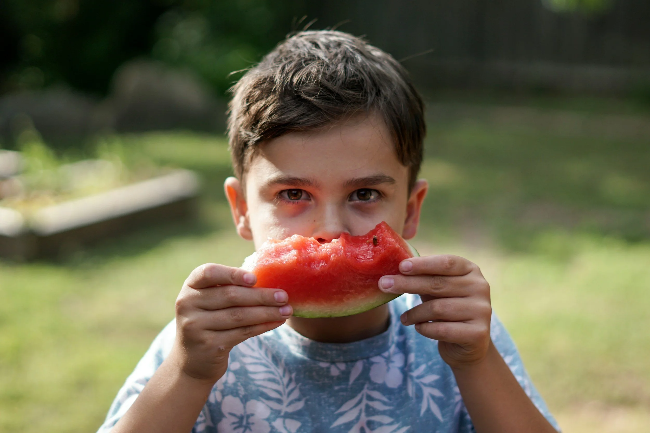 Young boy with brown hair and light skin eating a slice of watermelon outdoors.