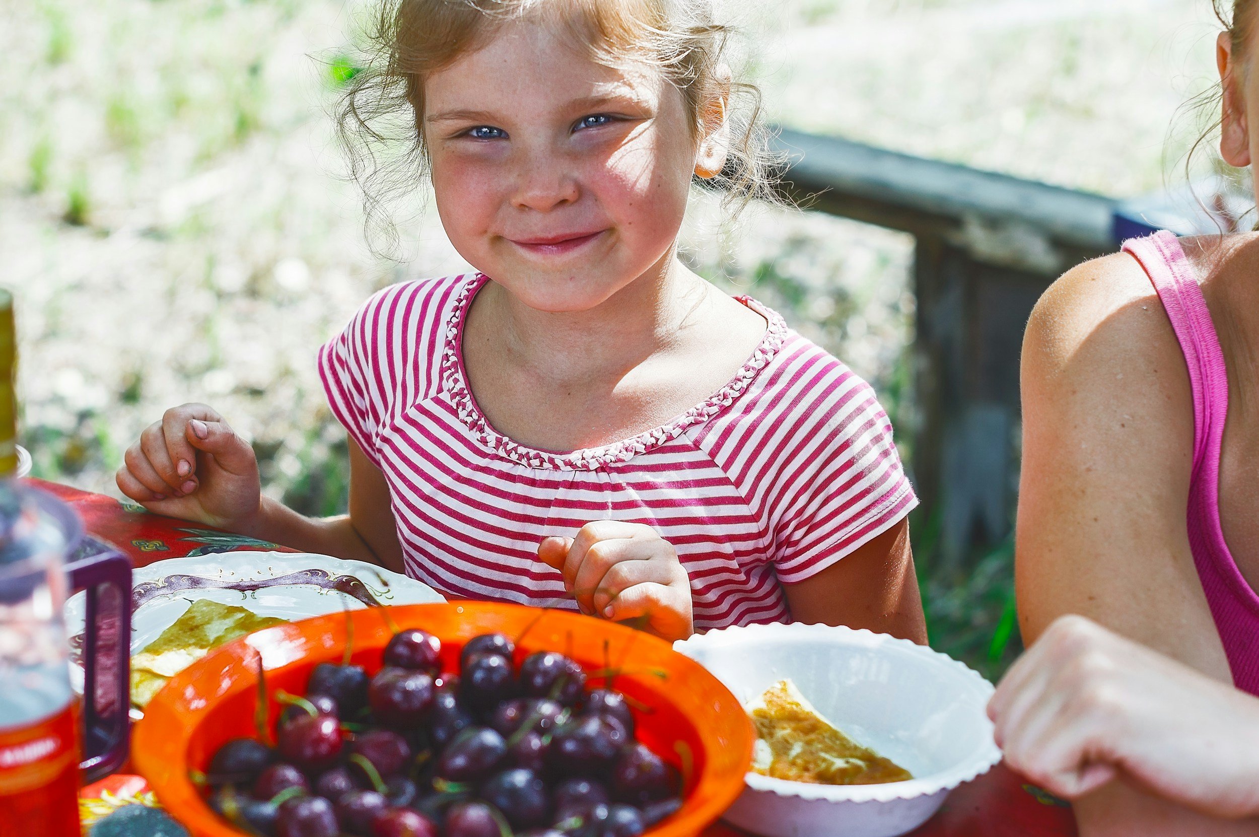 A young girl with curly hair and a striped pink and white shirt smiling at the camera while sitting at an outdoor table with a bowl of cherries and pie.