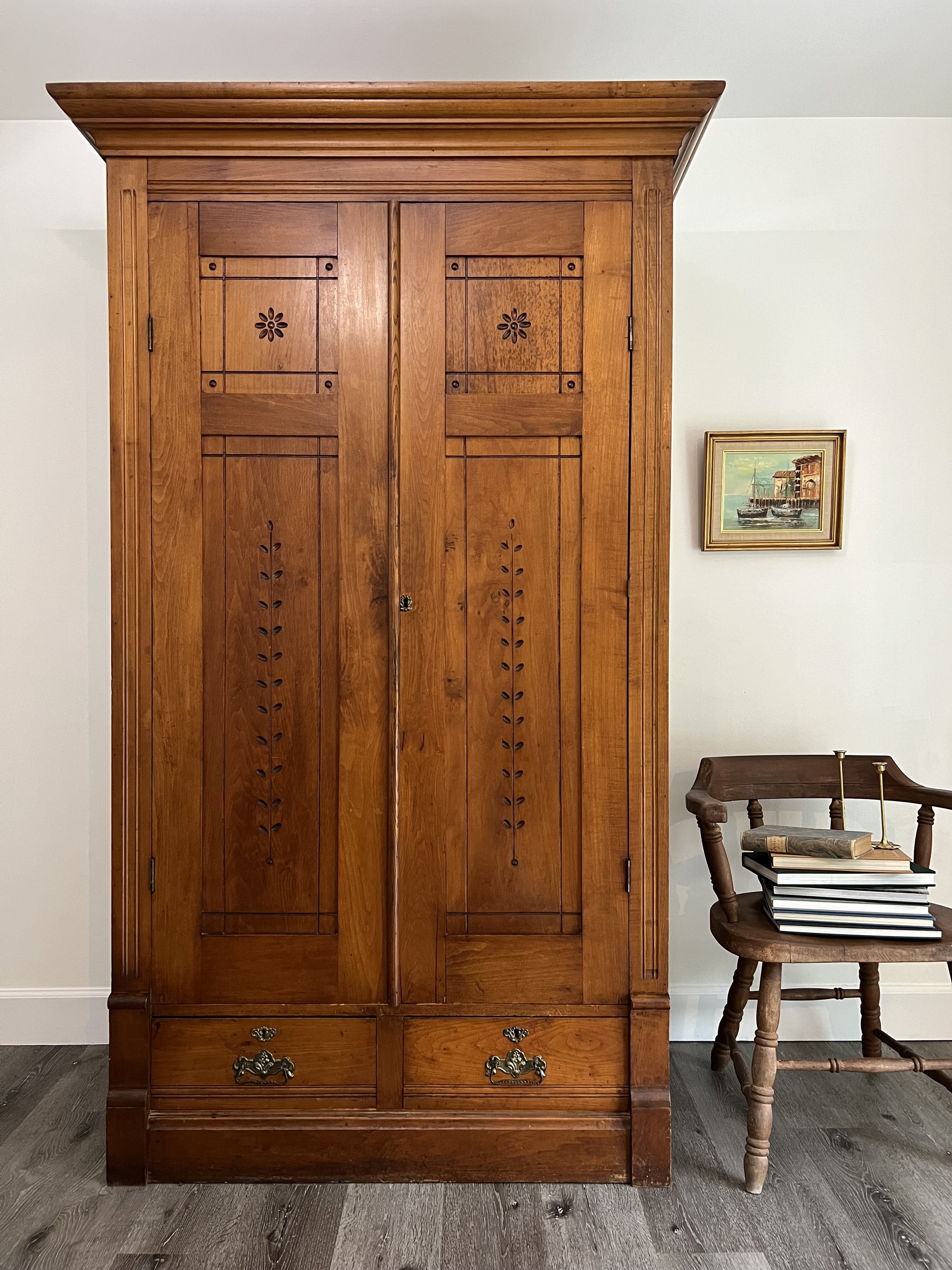 A tall wooden armoire with decorative panels and brass handles, positioned against a white wall, next to a small wooden chair with a stack of books on it, in a room with wood flooring.
