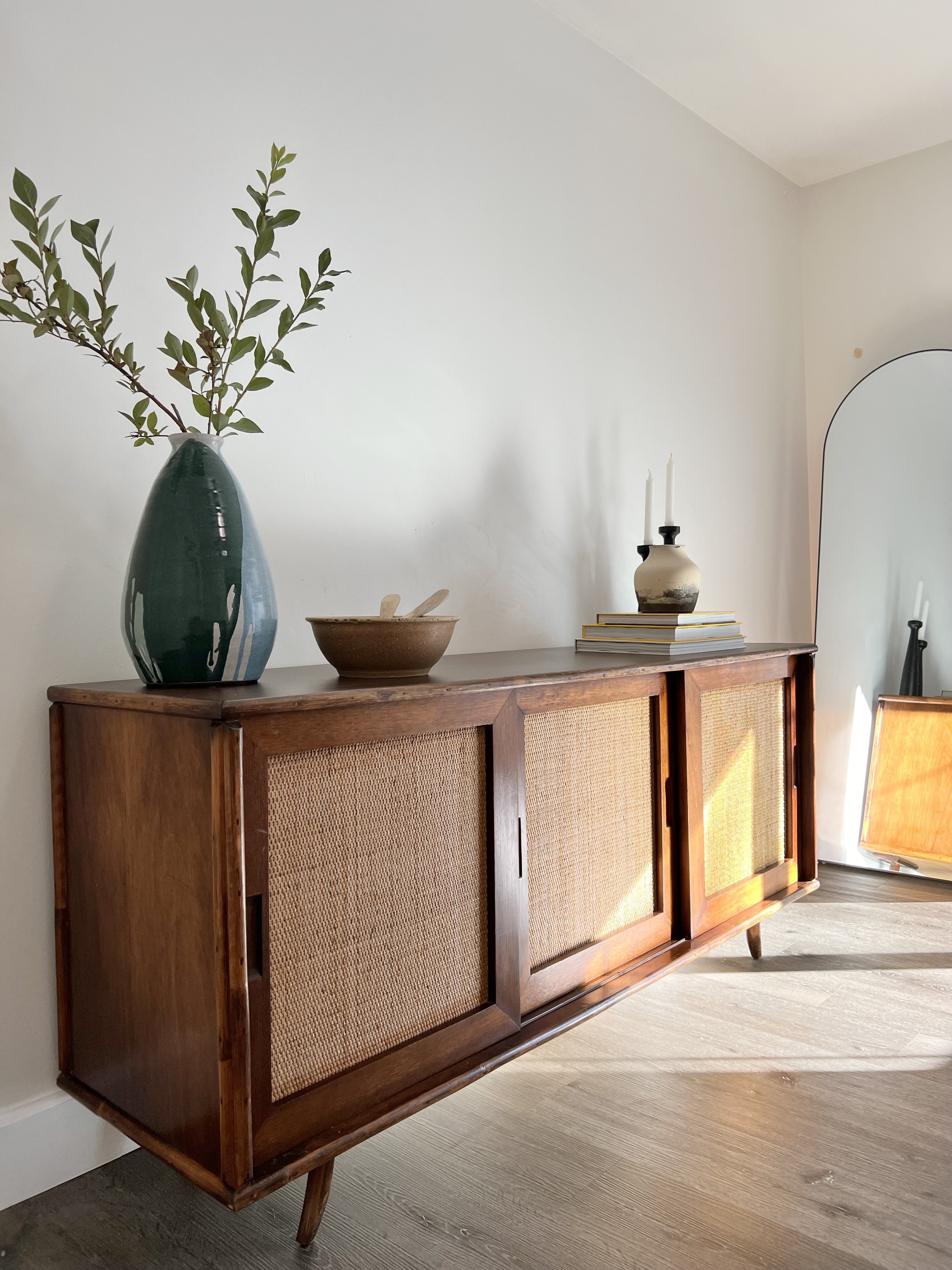 A wooden sideboard with cane sliding doors, topped with a large green vase with branches, a brown ceramic bowl, and a smaller vase with candles on top. A white and black object is leaning against the wall in the background.