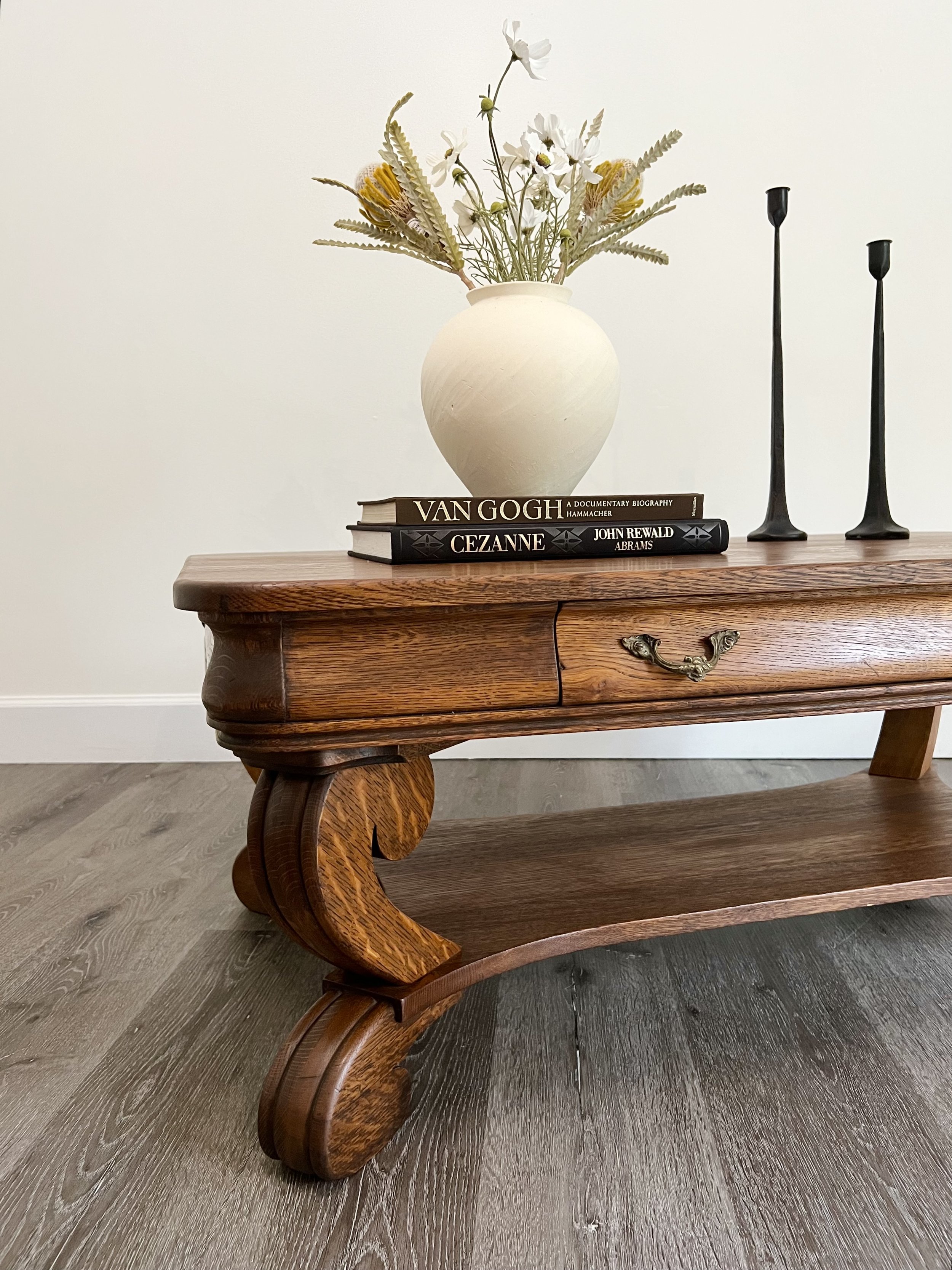 A vintage wooden coffee table with carved legs, topped with a white vase containing white and yellow flowers, two books titled 'Van Gogh' and 'Cezanne', and two tall black candles.