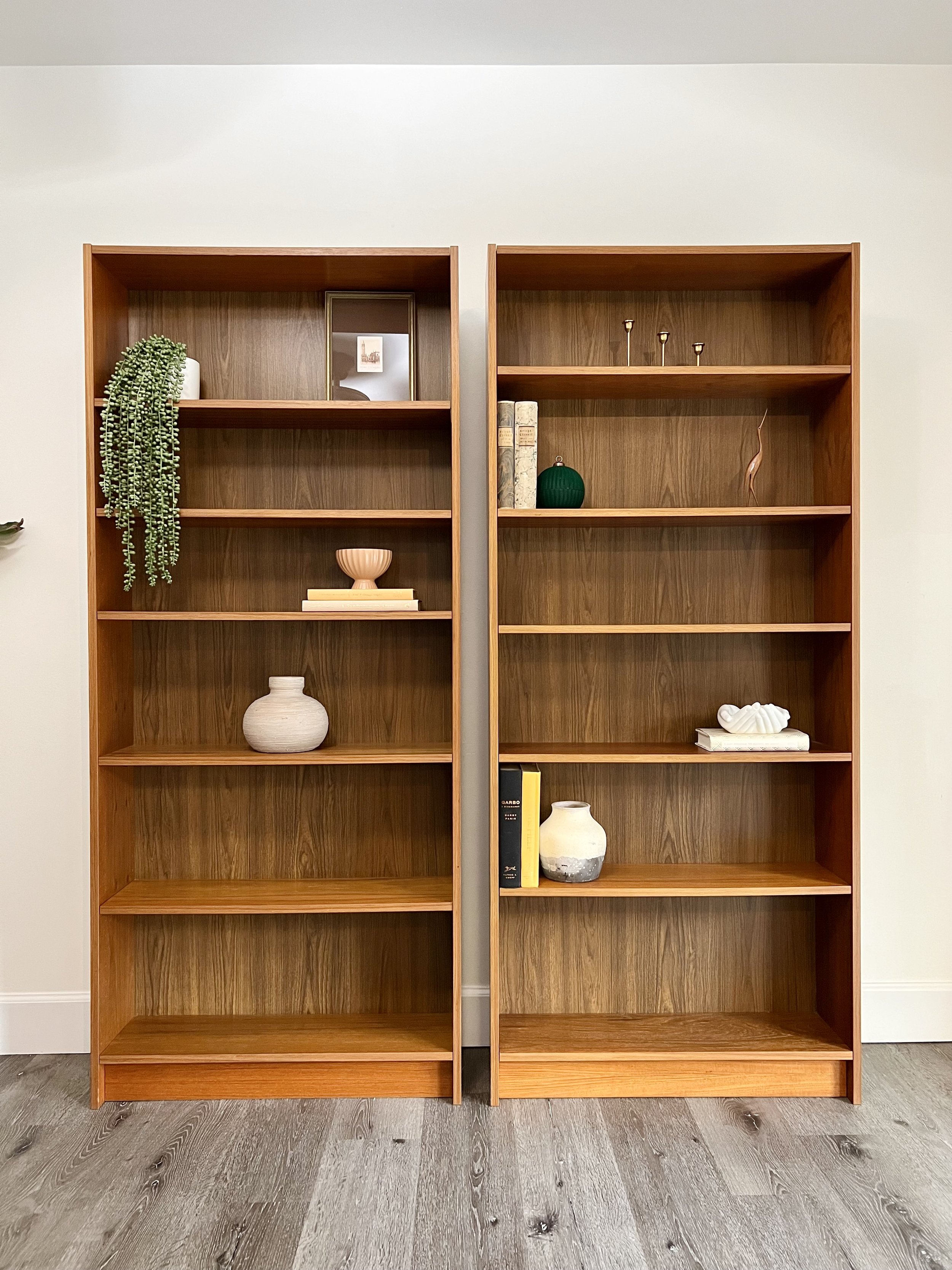 Two wooden bookshelves against a plain wall, decorated with vases, books, and small decorative items, located on a gray wooden floor.