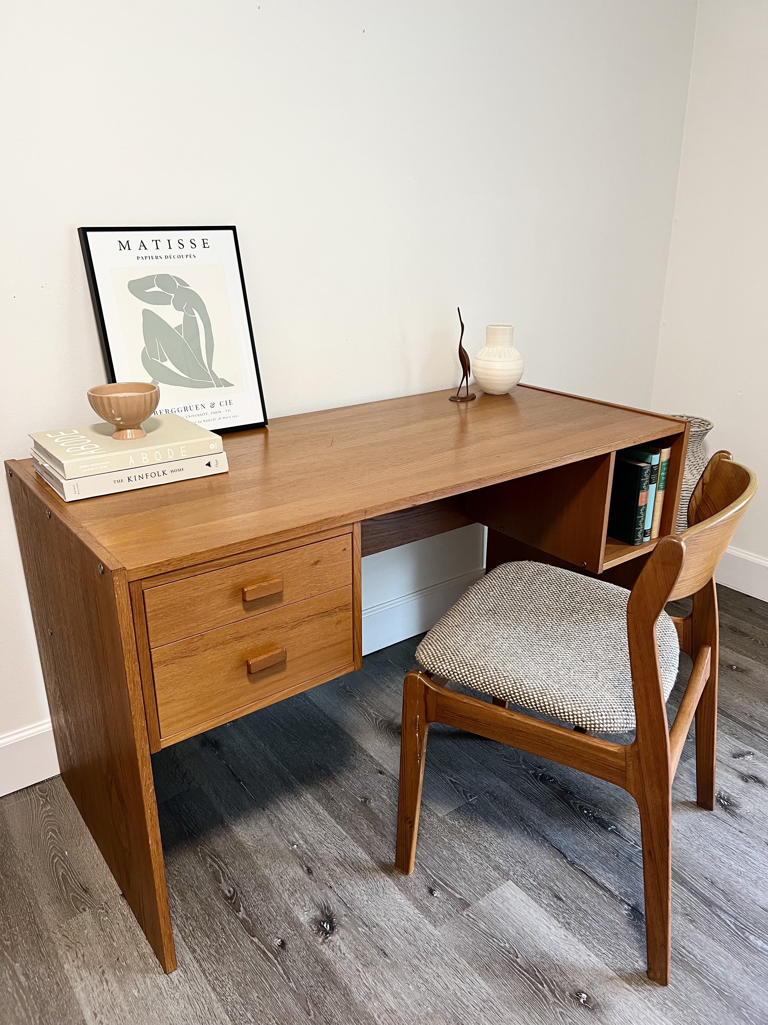Mid-century wooden desk with two drawers and a light-colored upholstered chair. Decor on the desk includes a framed Matisse art print, stacked books, a small ceramic bowl, a white vase, and a bronze sculpture of a figure. The desk is against a plain
