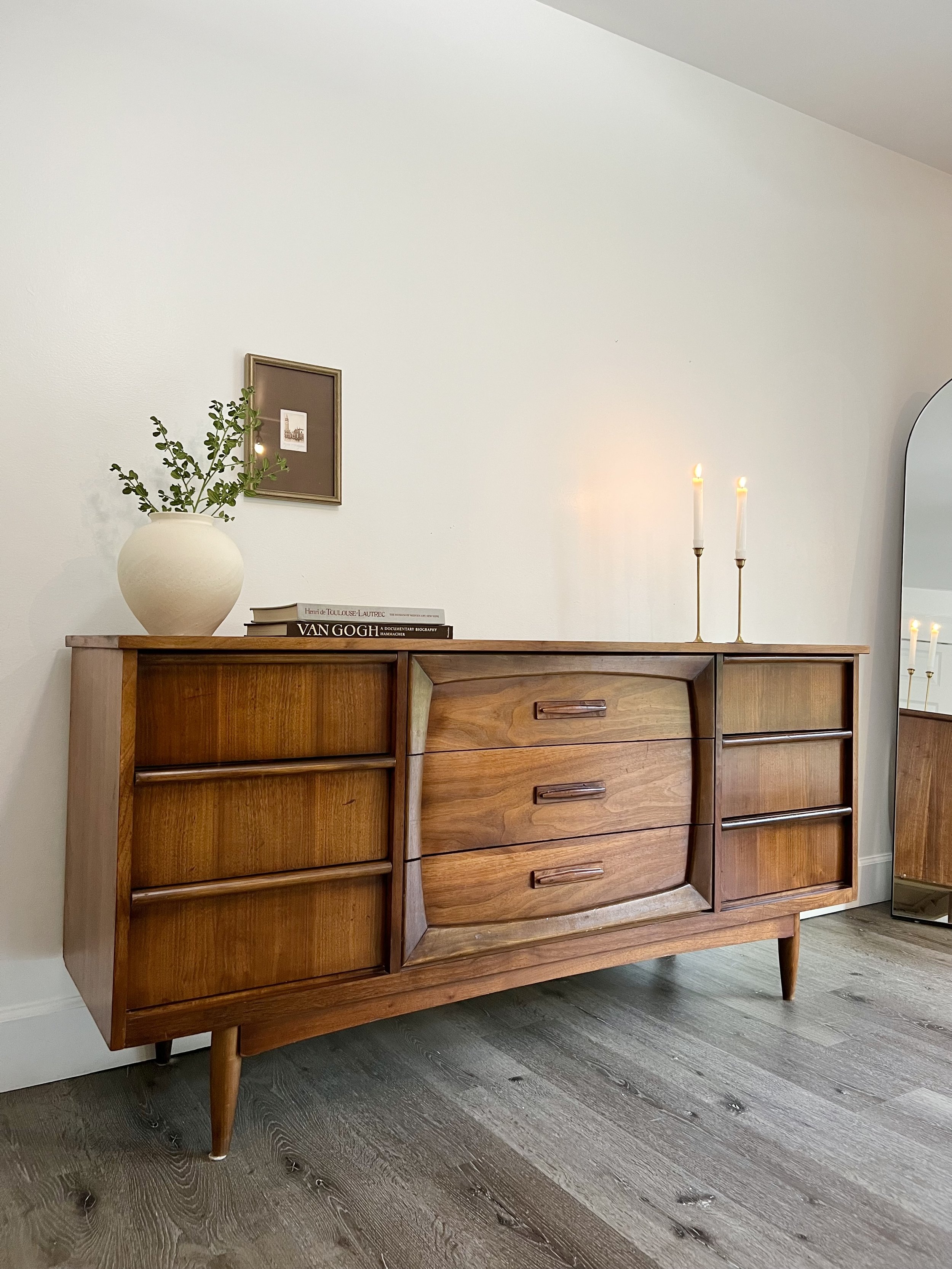 Mid-century wooden sideboard with books, a vase with greenery, and lit candles on top, standing against a plain white wall with a small framed artwork.