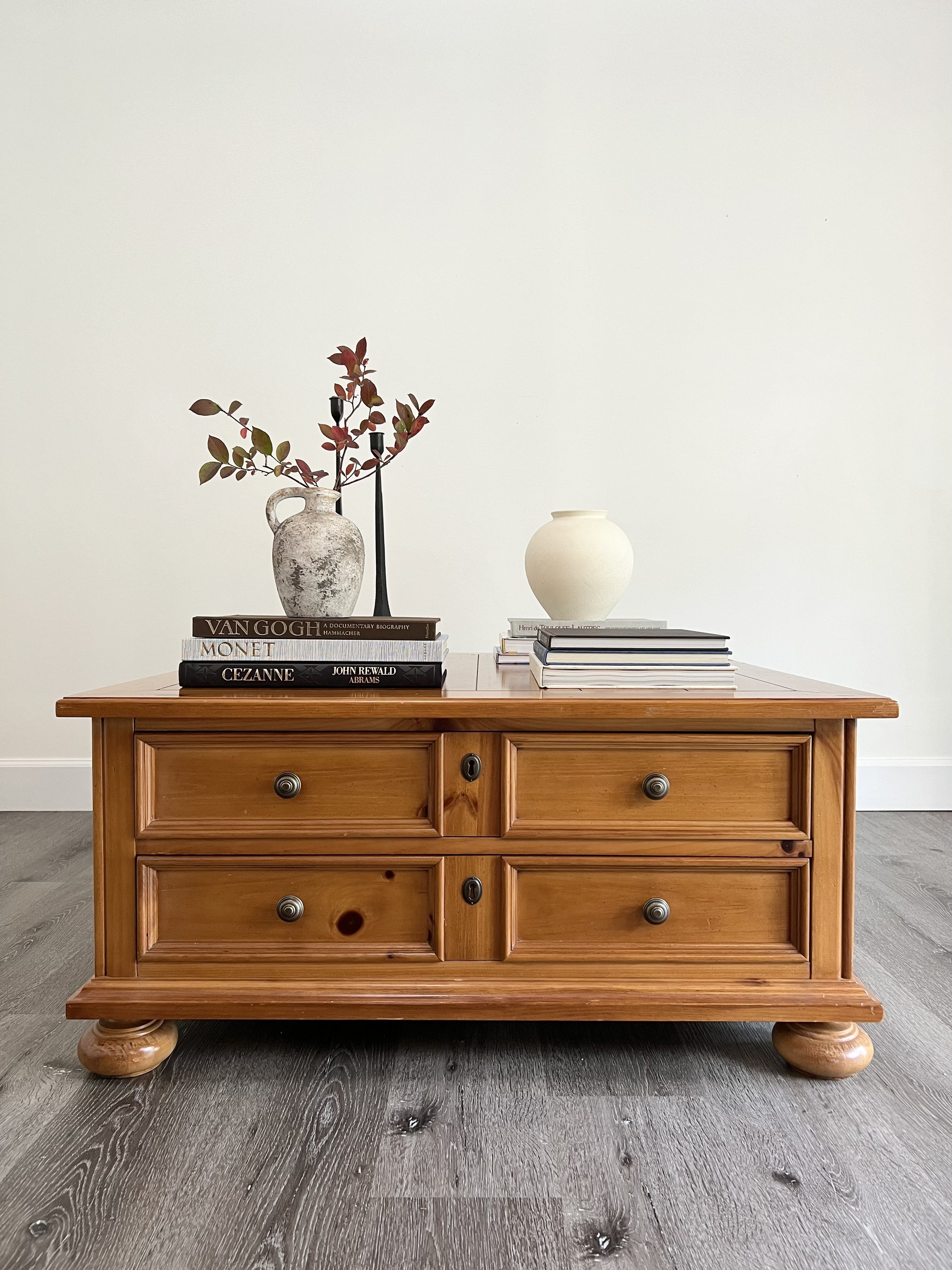 A wooden dresser with four drawers, decorated with books, vases, and a plant, against a plain light-colored wall and gray flooring.