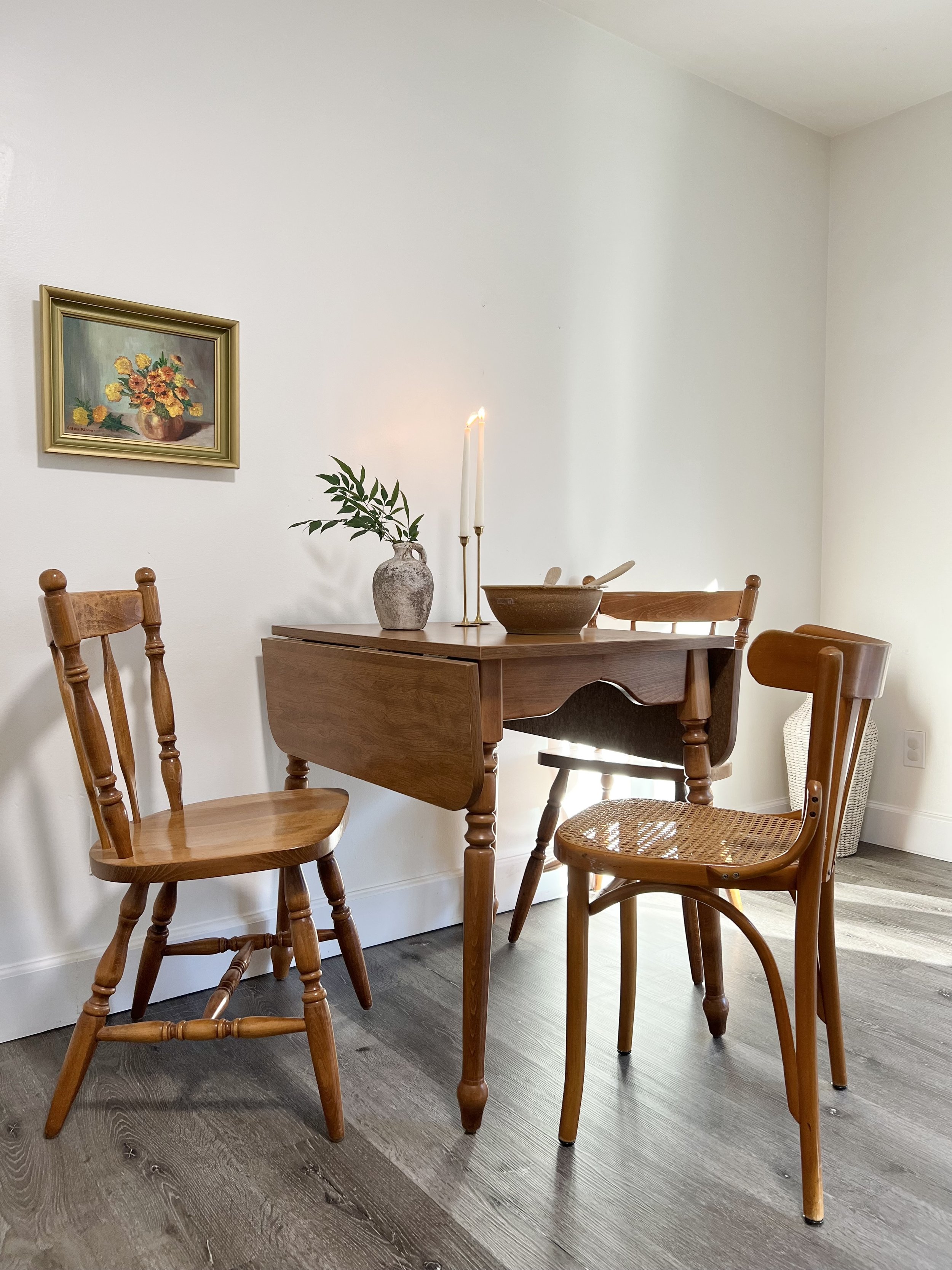 A cozy dining area with a wooden table, two wooden chairs, a white wall, a framed floral painting, and a small plant in a vase, with soft natural light.