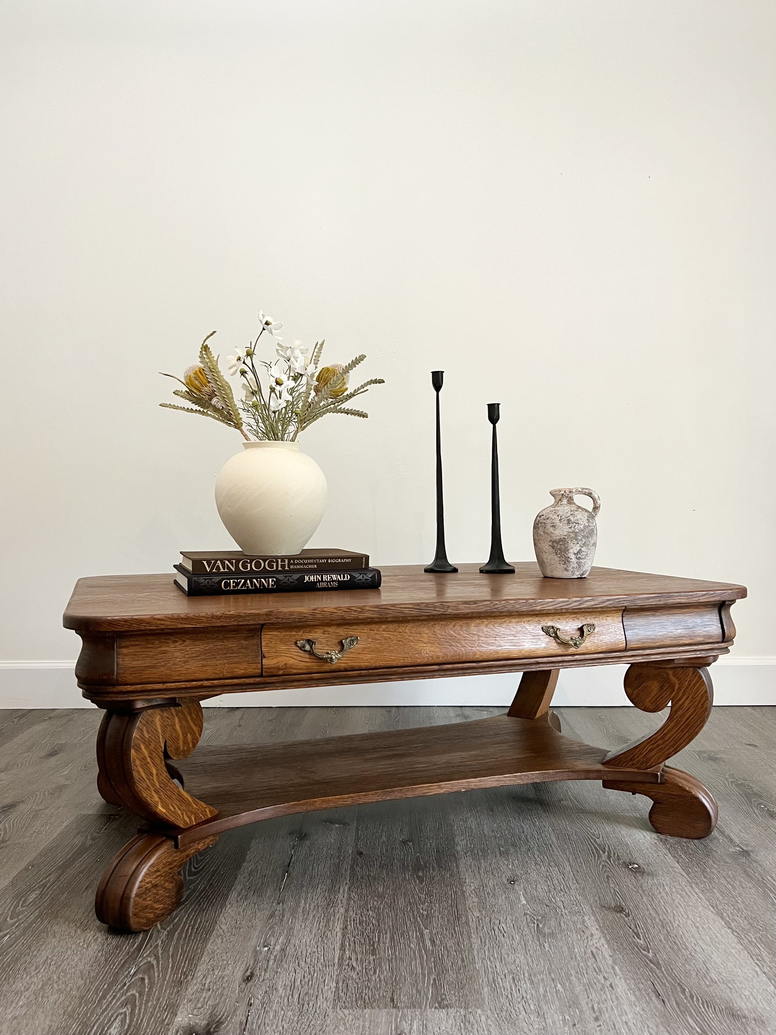 A wooden coffee table decorated with a vase of flowers, two black candlesticks, a small ceramic pitcher, and two books against a plain white wall.