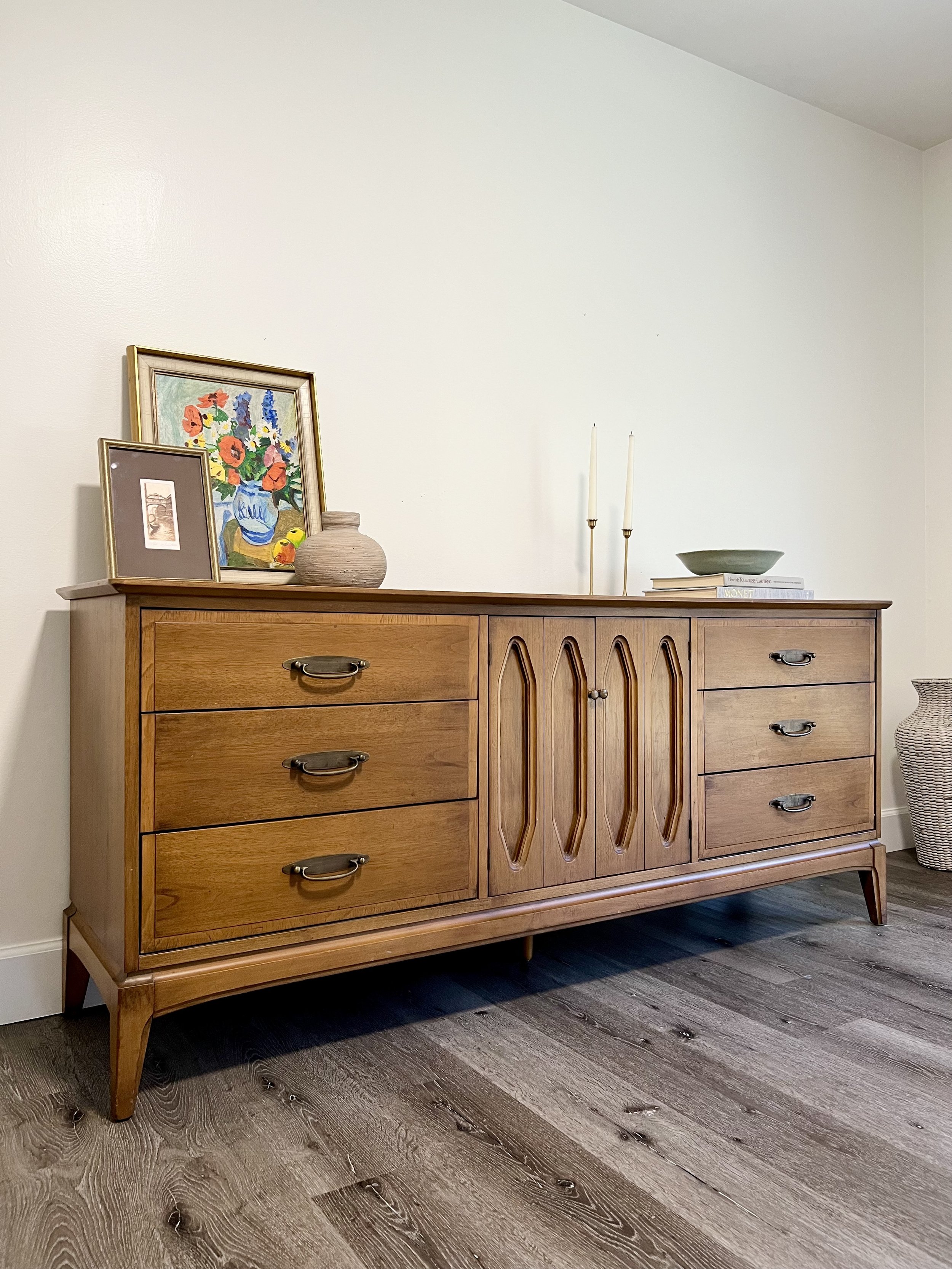 A mid-century modern wooden sideboard with drawers and cabinet doors, situated against a white wall, decorated with framed artwork, a vase, candlesticks, and bowls on top, with a woven basket on the floor nearby.