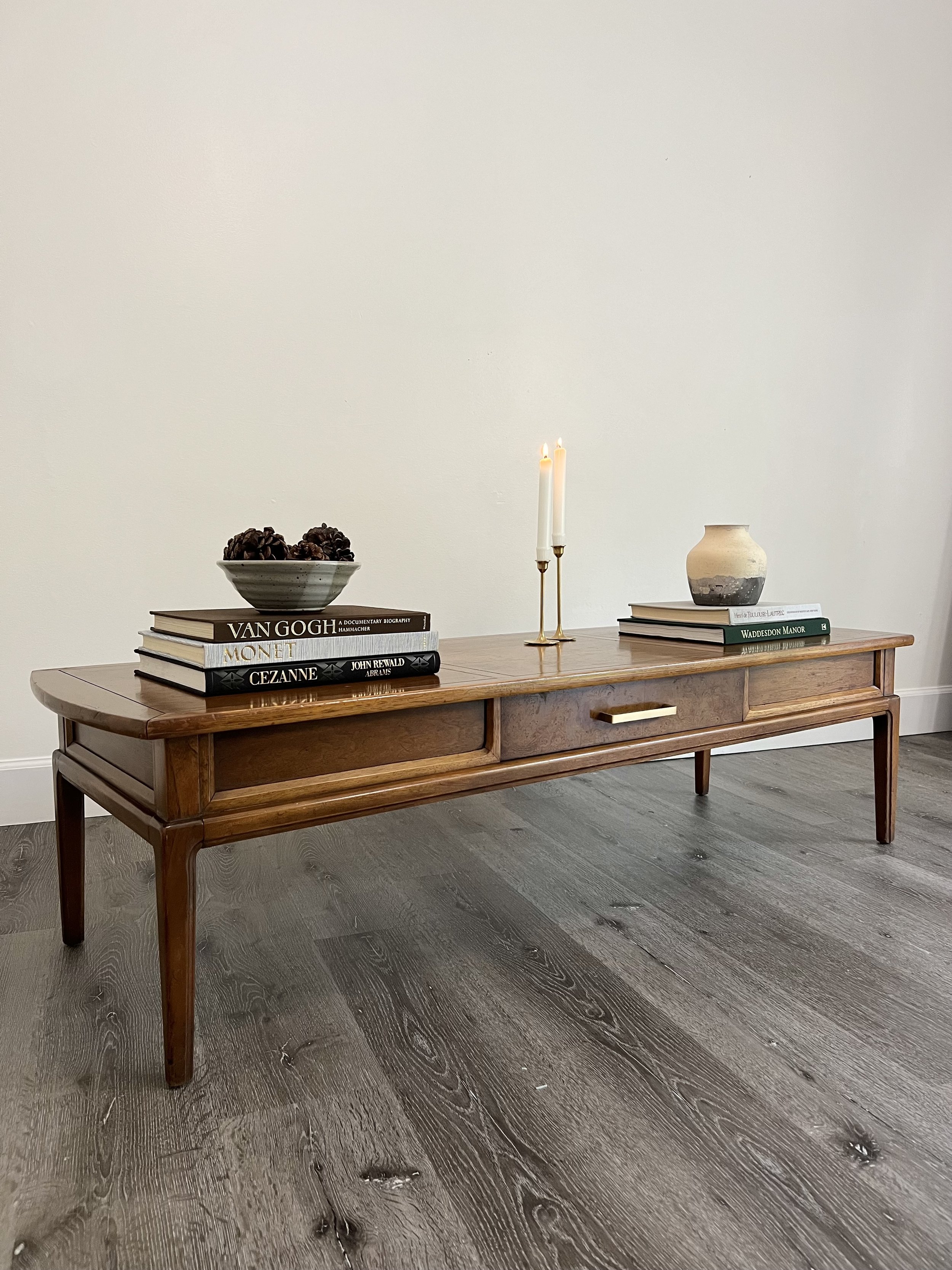 Wooden console table with a stack of books, a bowl of pinecones, a pair of lit candles, and a ceramic vase with a stack of books beside it, set against a plain white wall.