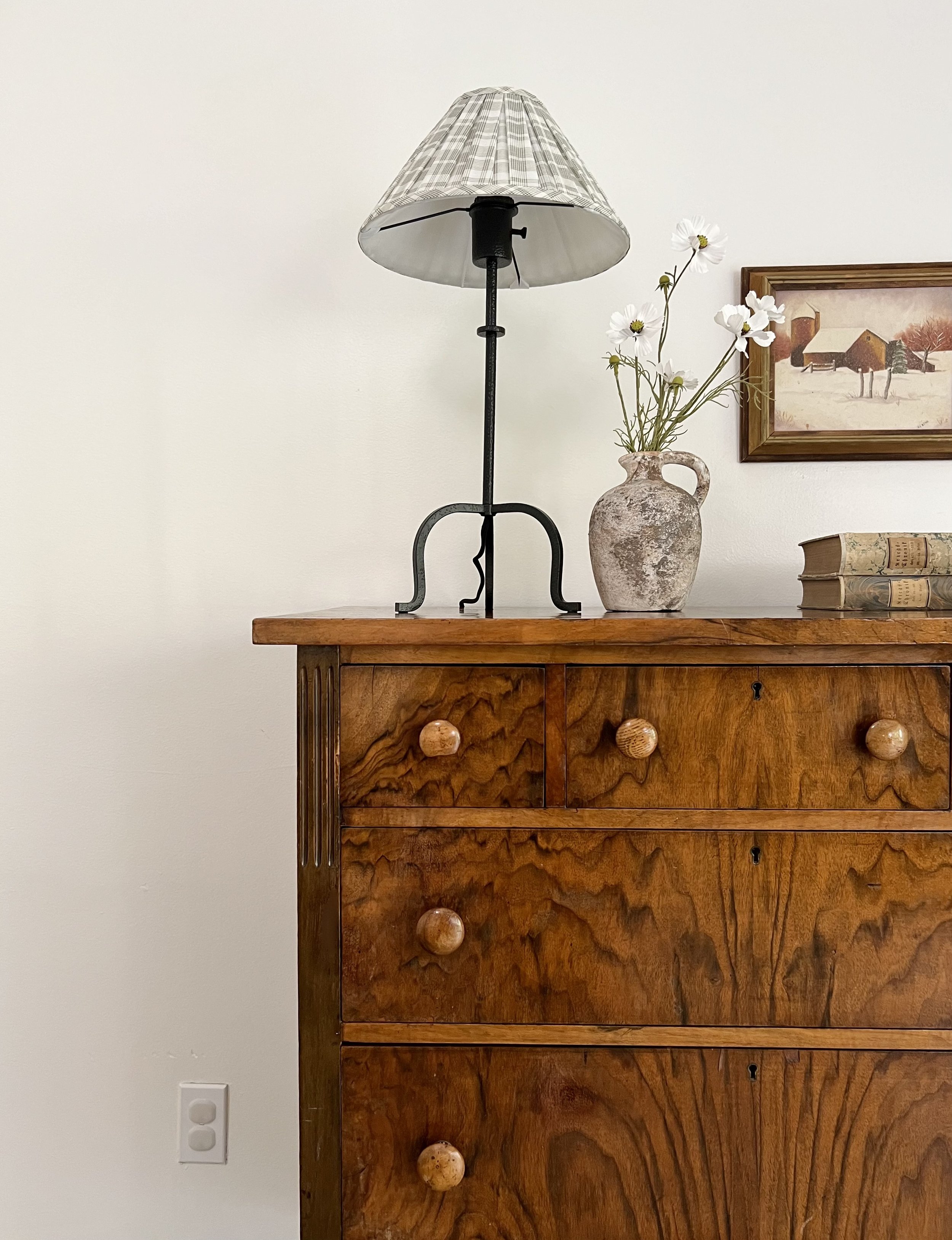 A wooden dresser with a tabletop lamp, a ceramic vase with flowers, and a framed painting hanging on a white wall.