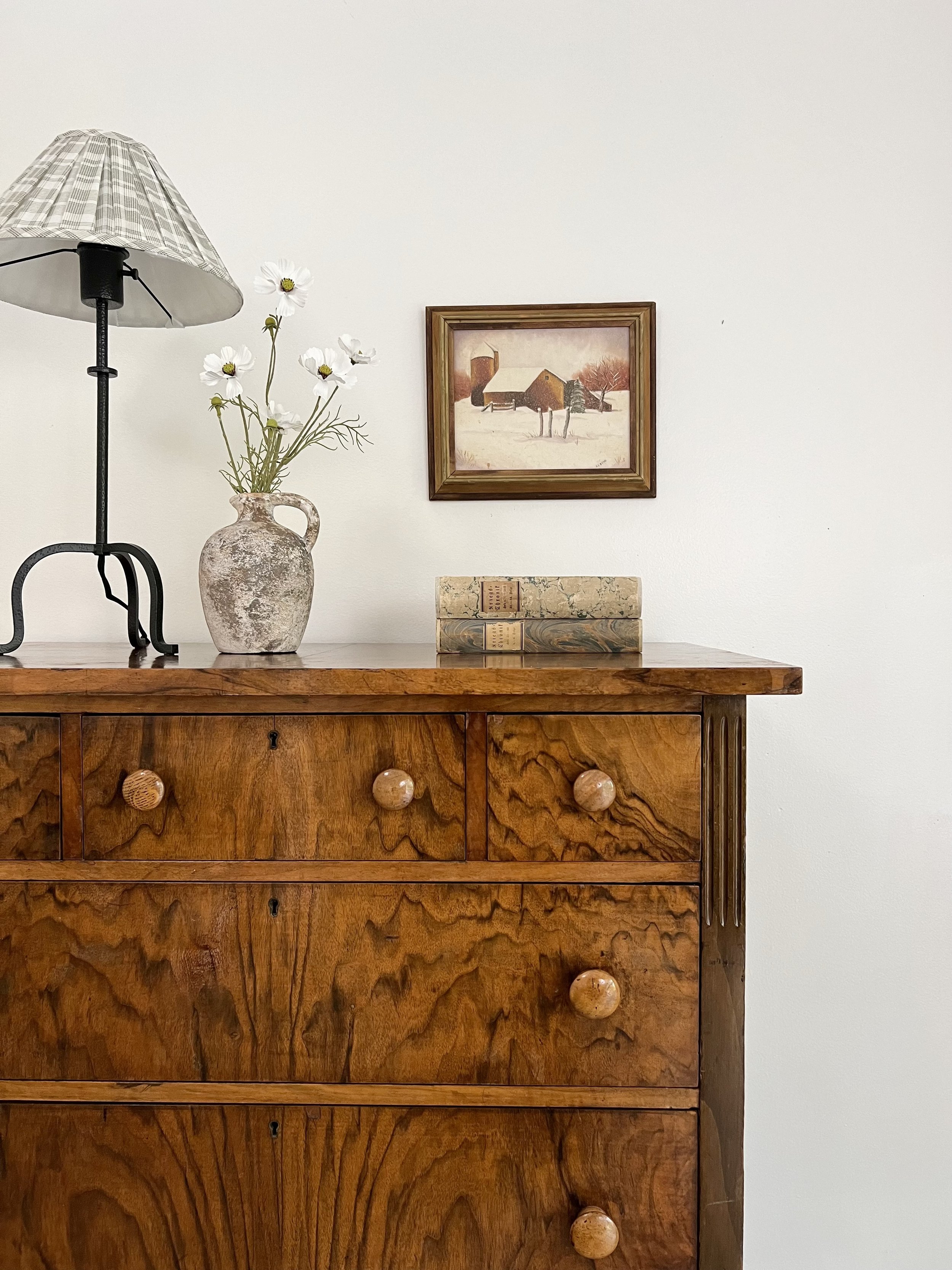 A wooden dresser with round knobs, a lamp, a ceramic vase with white flowers, a set of books, a painting of a rural winter scene, and a white wall.