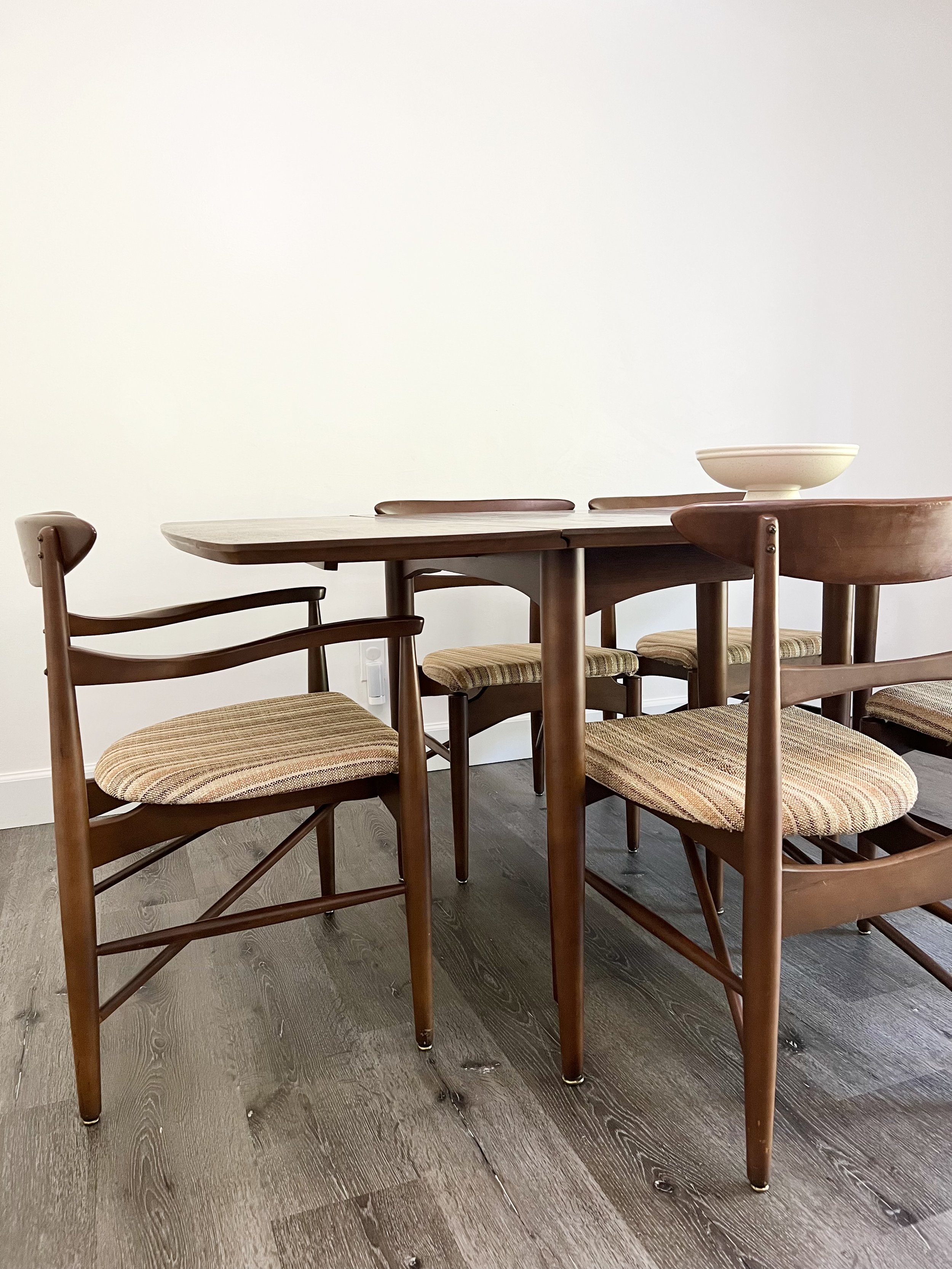 A wooden dining table with four matching chairs, one with a striped upholstered seat, and a white bowl on top, against a plain white wall and dark wood floor.