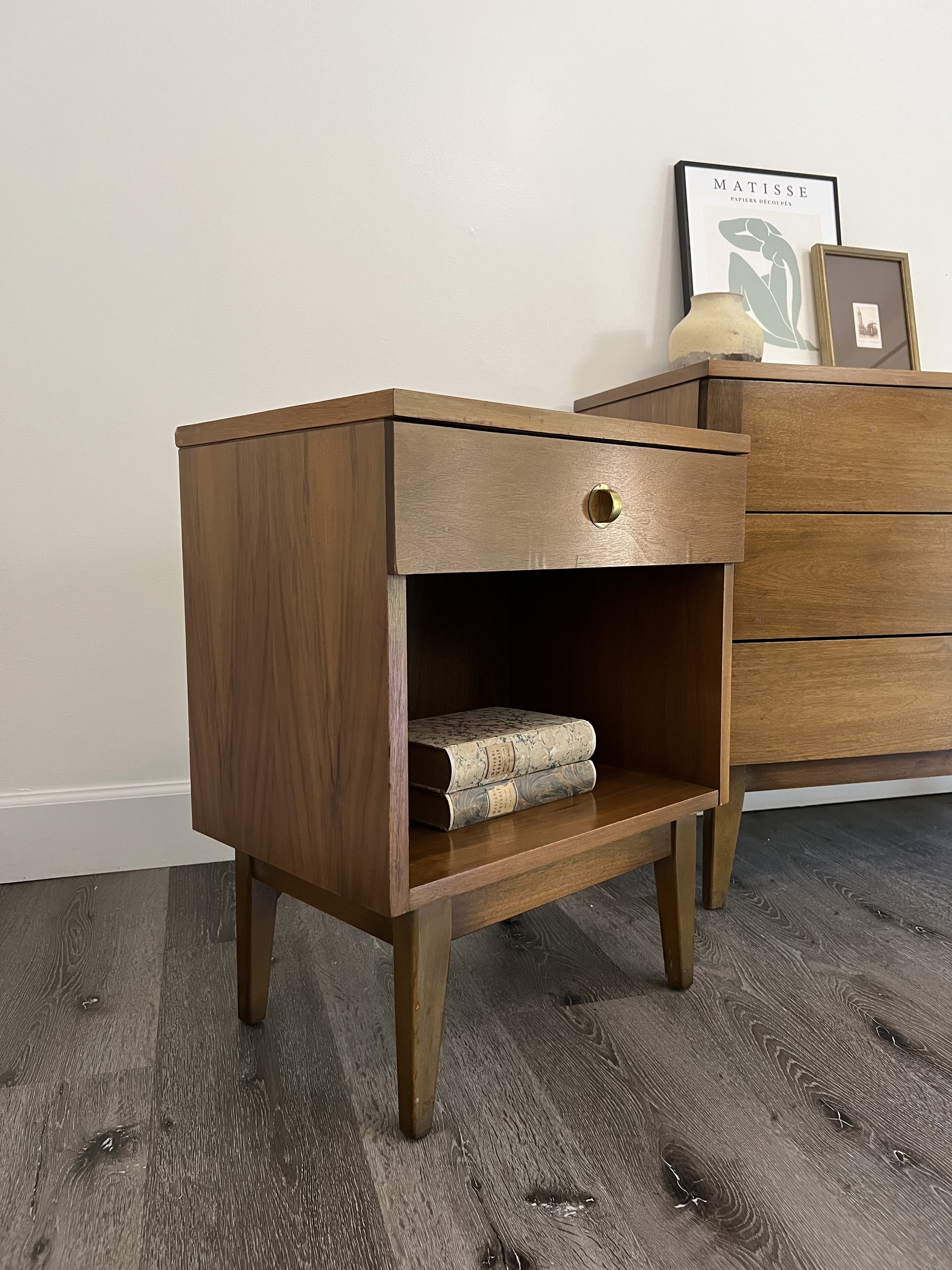 A mid-century modern wooden nightstand with two drawers and four tapered legs, holding three vintage-style books. In the background, there is a wooden dresser with framed artwork and a small vase on top.