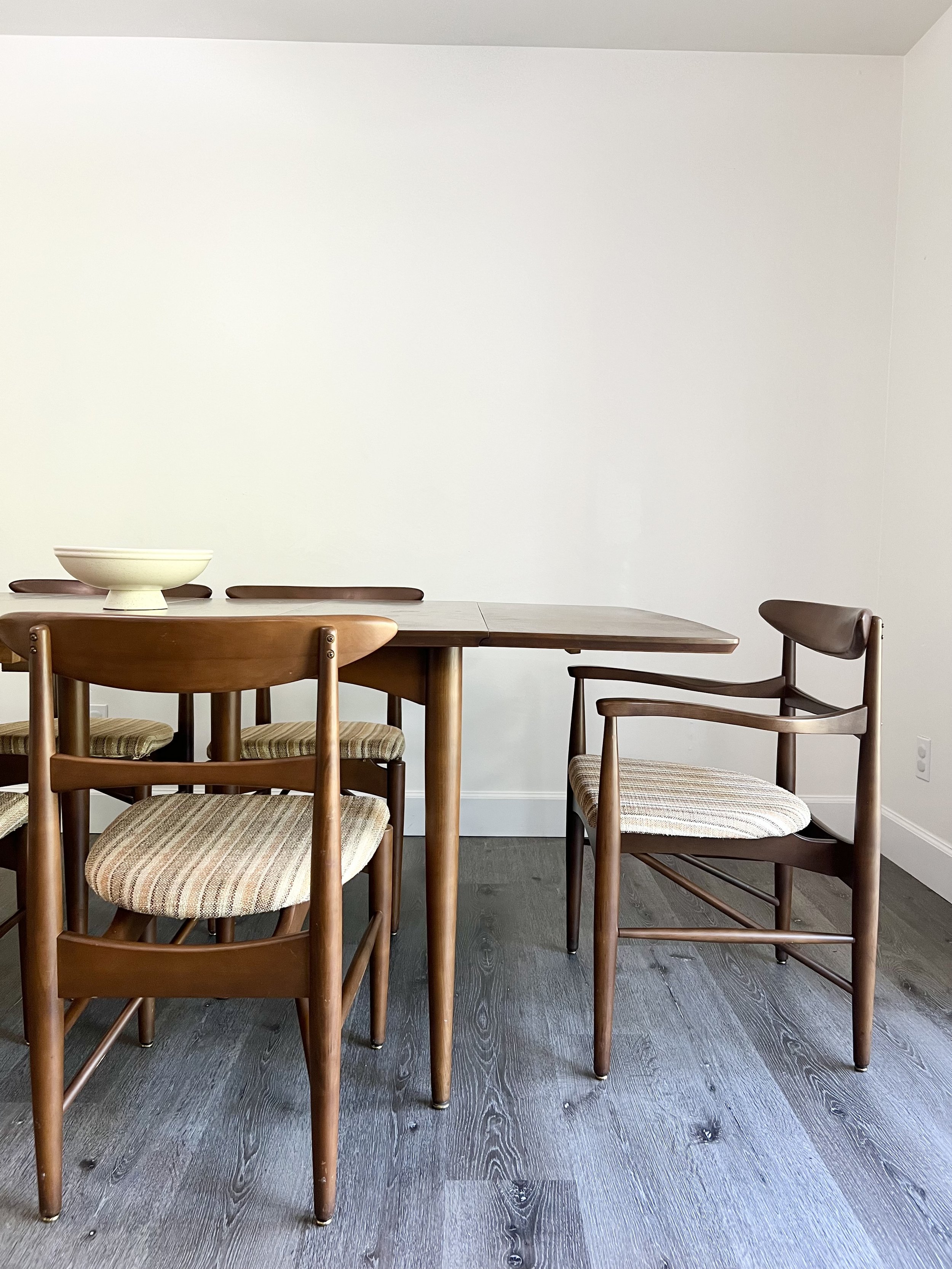 A wooden dining table with several chairs, two of which have cushioned seats with striped fabric. There is a large, decorative bowl on the table, and the room has light-colored walls and dark hardwood floors.