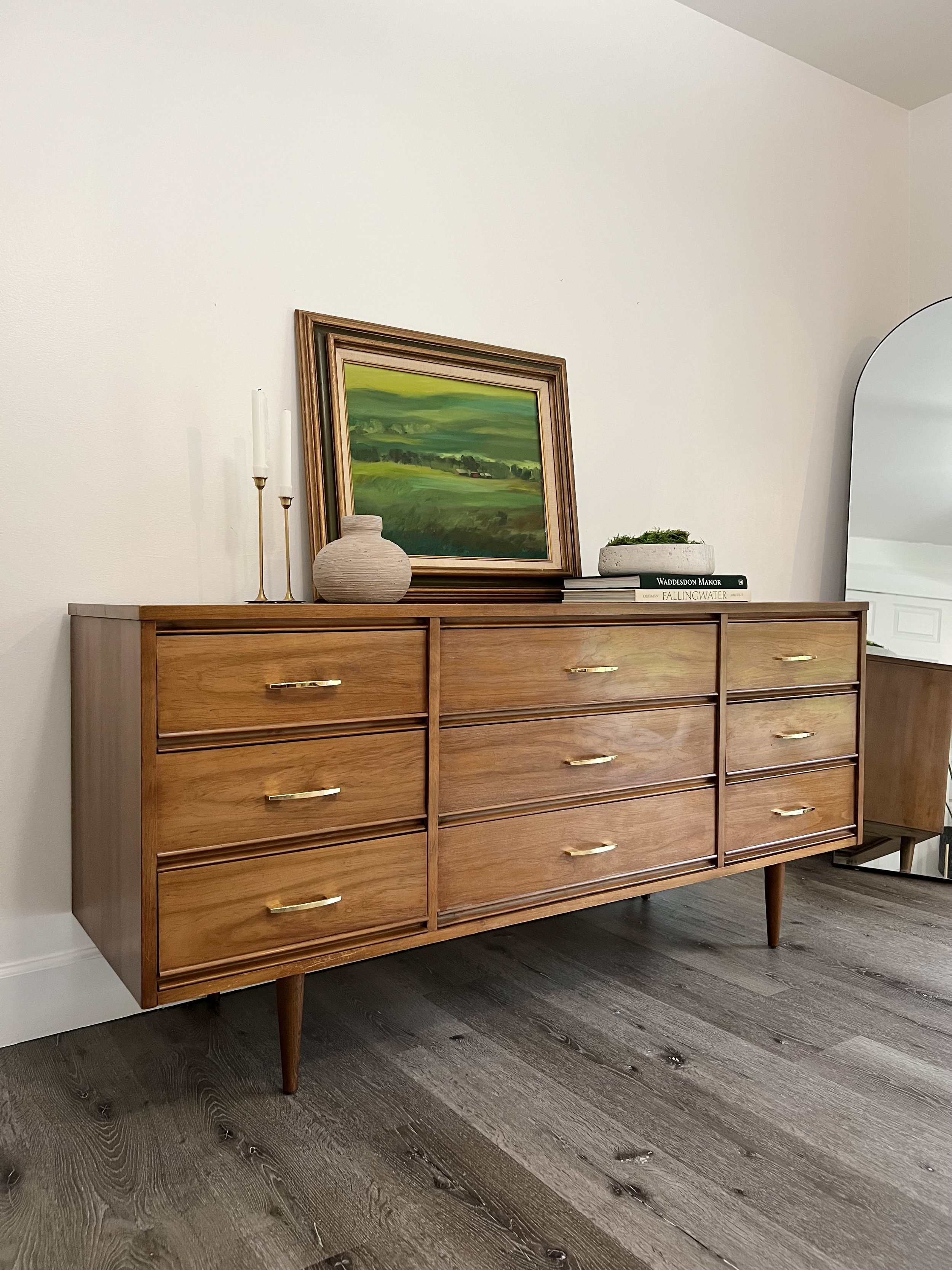 A mid-century modern wooden dresser with six drawers and gold handles, topped with a framed landscape painting, three white candles on gold candlesticks, a small pottery vase, a stack of books, and a round concrete planter with greenery, next to a la