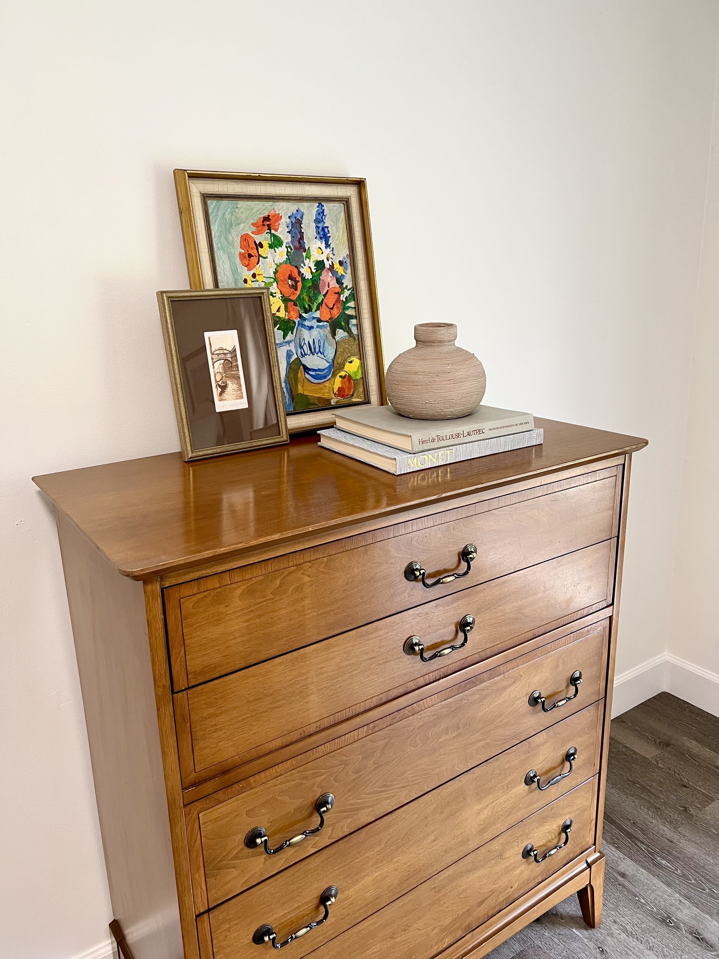 A wooden dresser with five drawers, decorated with framed art, books, a pottery vase, and a colorful floral painting on top.