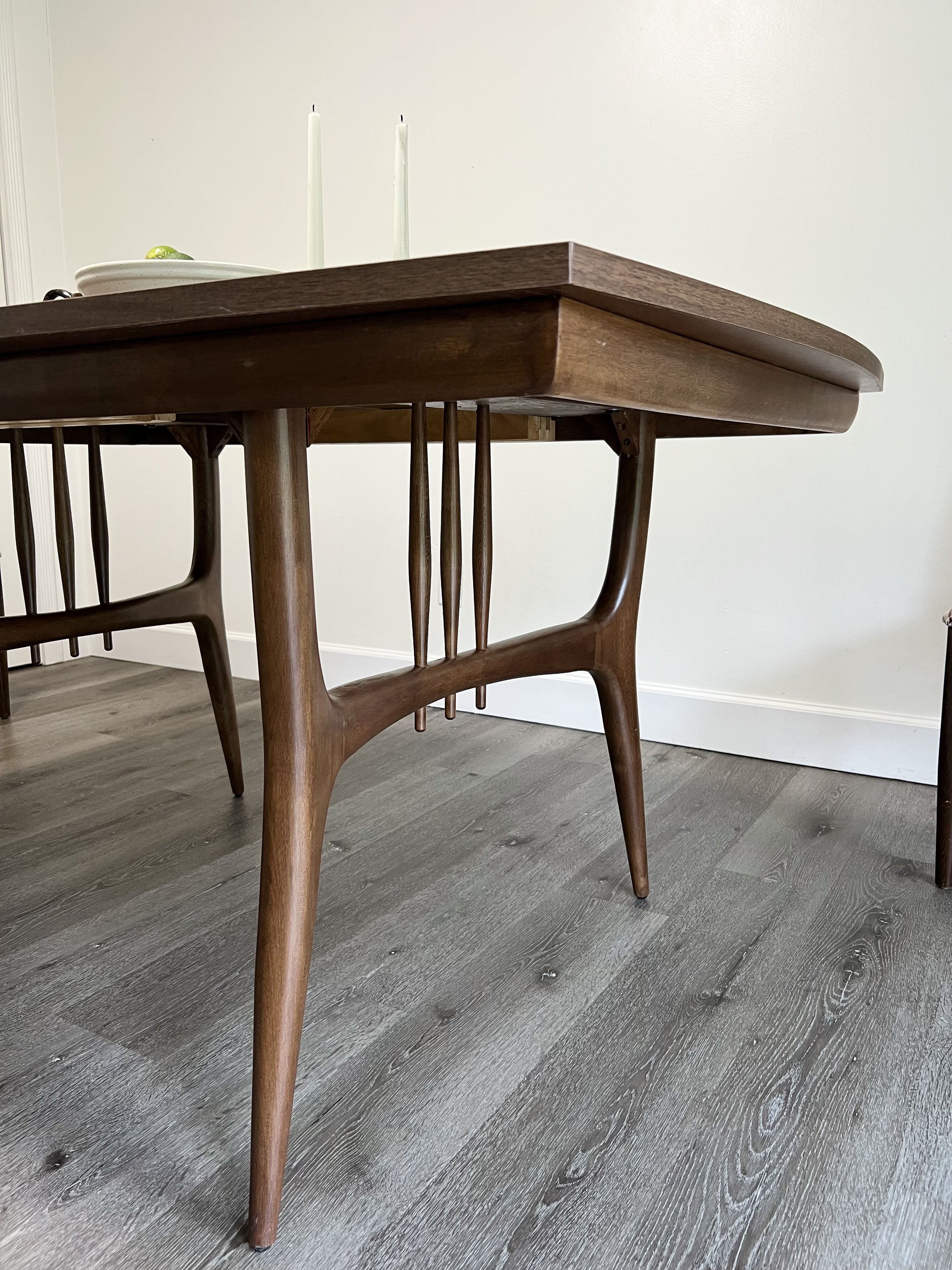 A wooden dining table with curved edges and matching chairs, situated on grey wood flooring against a plain light-colored wall.