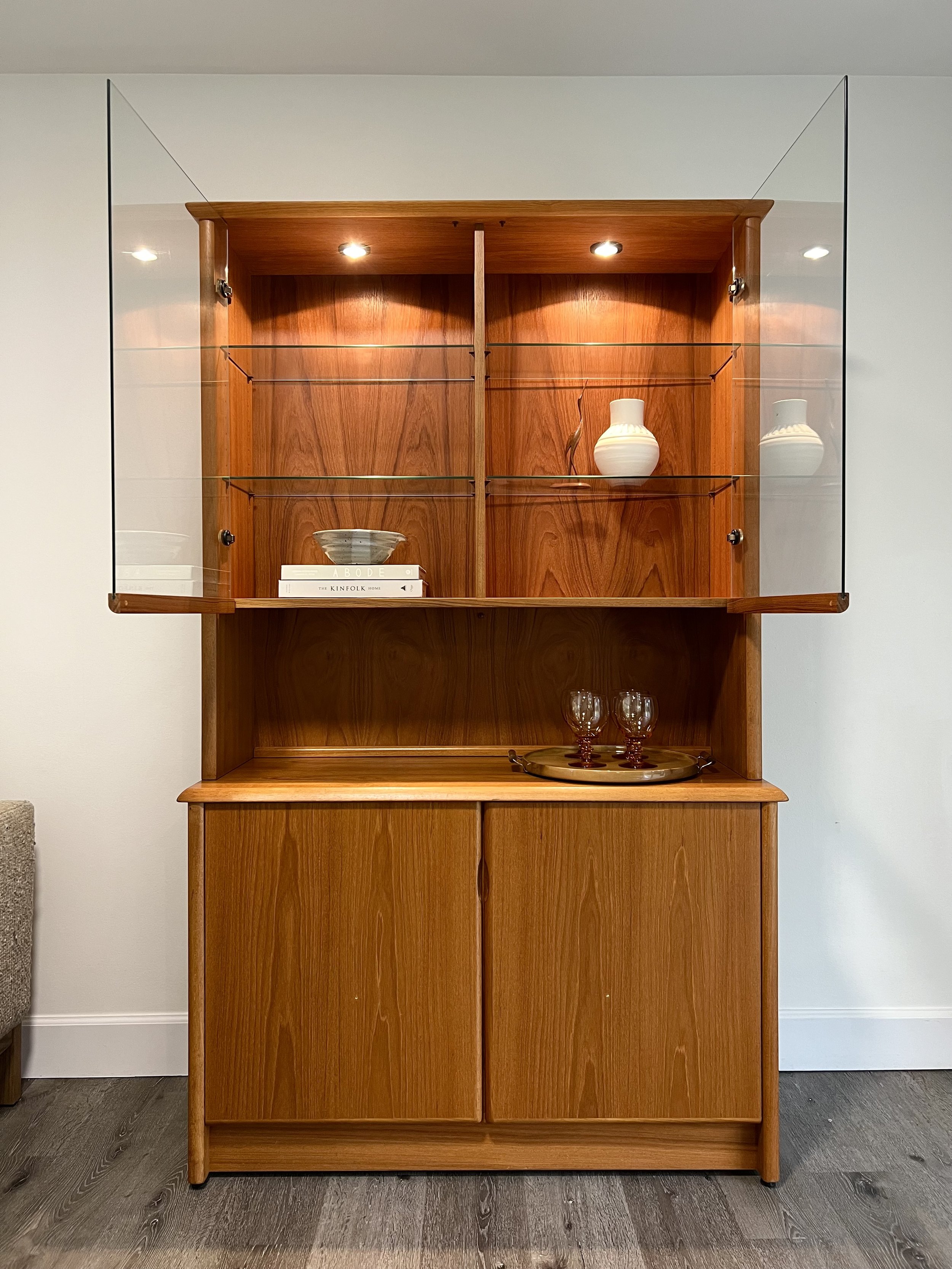 Mid-century modern wooden display cabinet with glass doors, containing decorative vases, bowls, books, and glassware, set against a white wall on a wooden floor.
