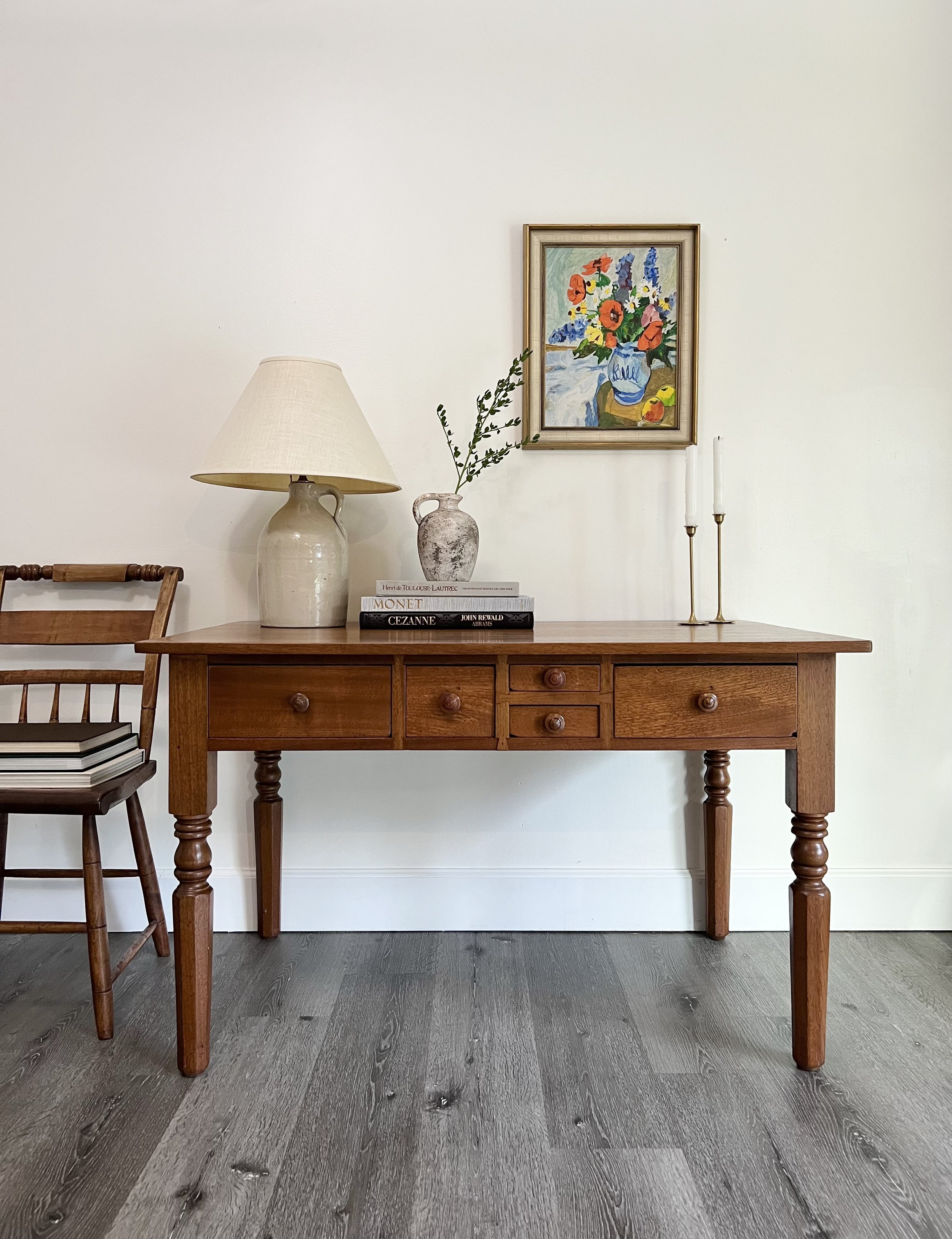 A wooden side table on a dark hardwood floor with a table lamp, decorative vases, stacked books, and tall candlesticks. A framed floral painting hangs on a white wall behind.