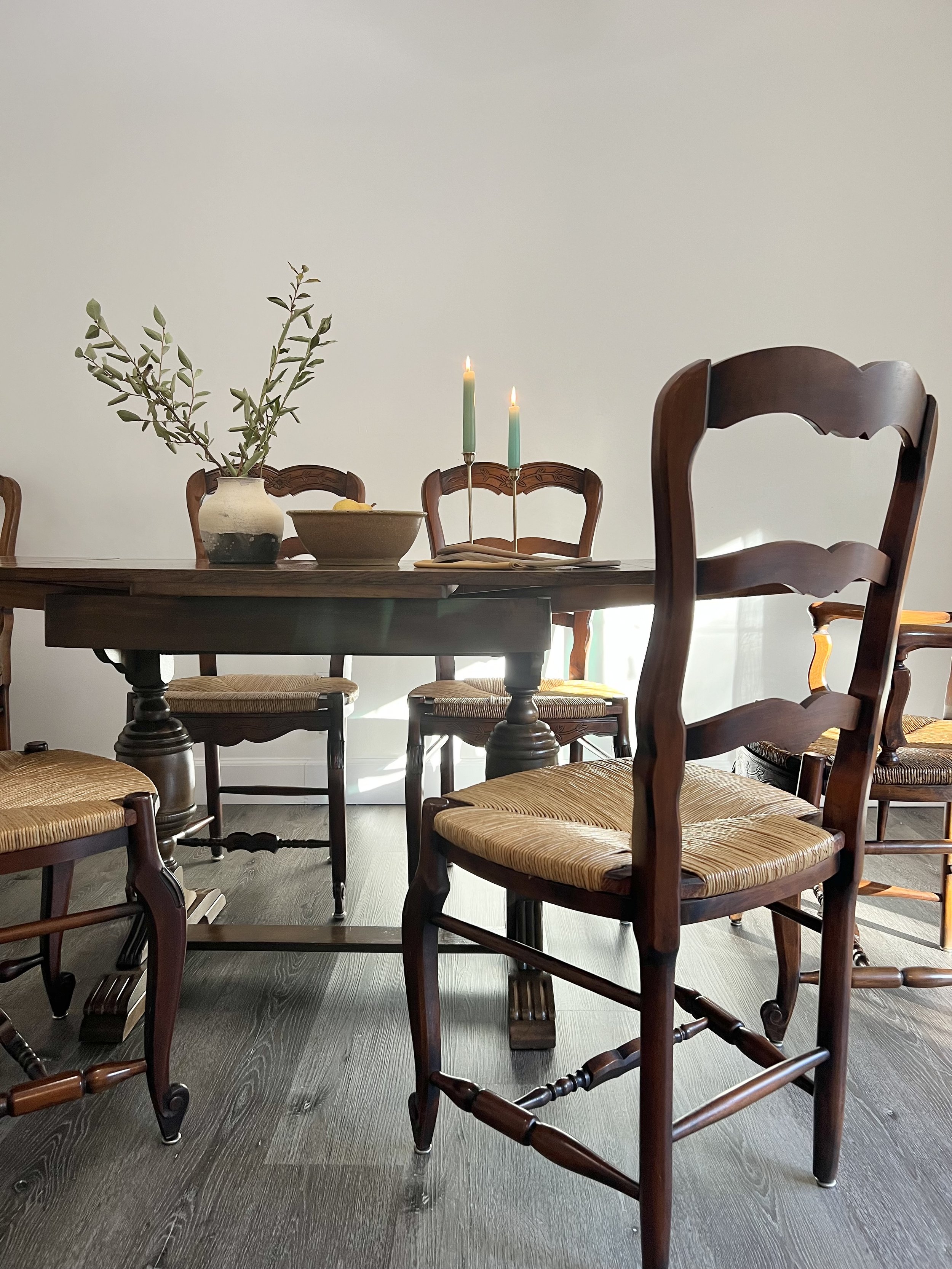 A wooden dining table with six matching wooden chairs, a ceramic vase with green leafy branches, a bowl with a lemon, and two lit green candles in holders on a white wall background.
