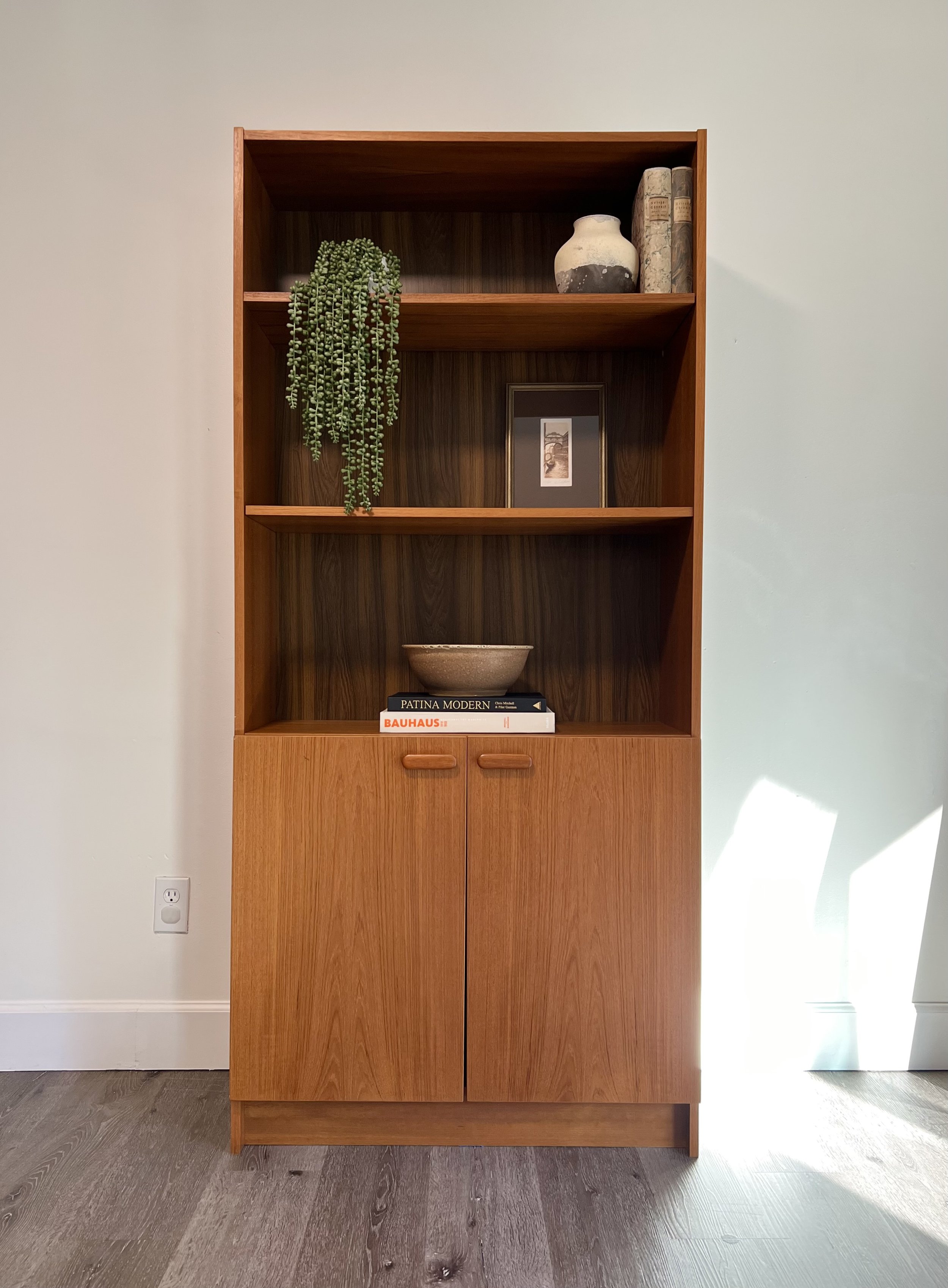 A wooden bookshelf with decor items and books, positioned against a plain white wall, with sunlight casting a shadow on the wall.