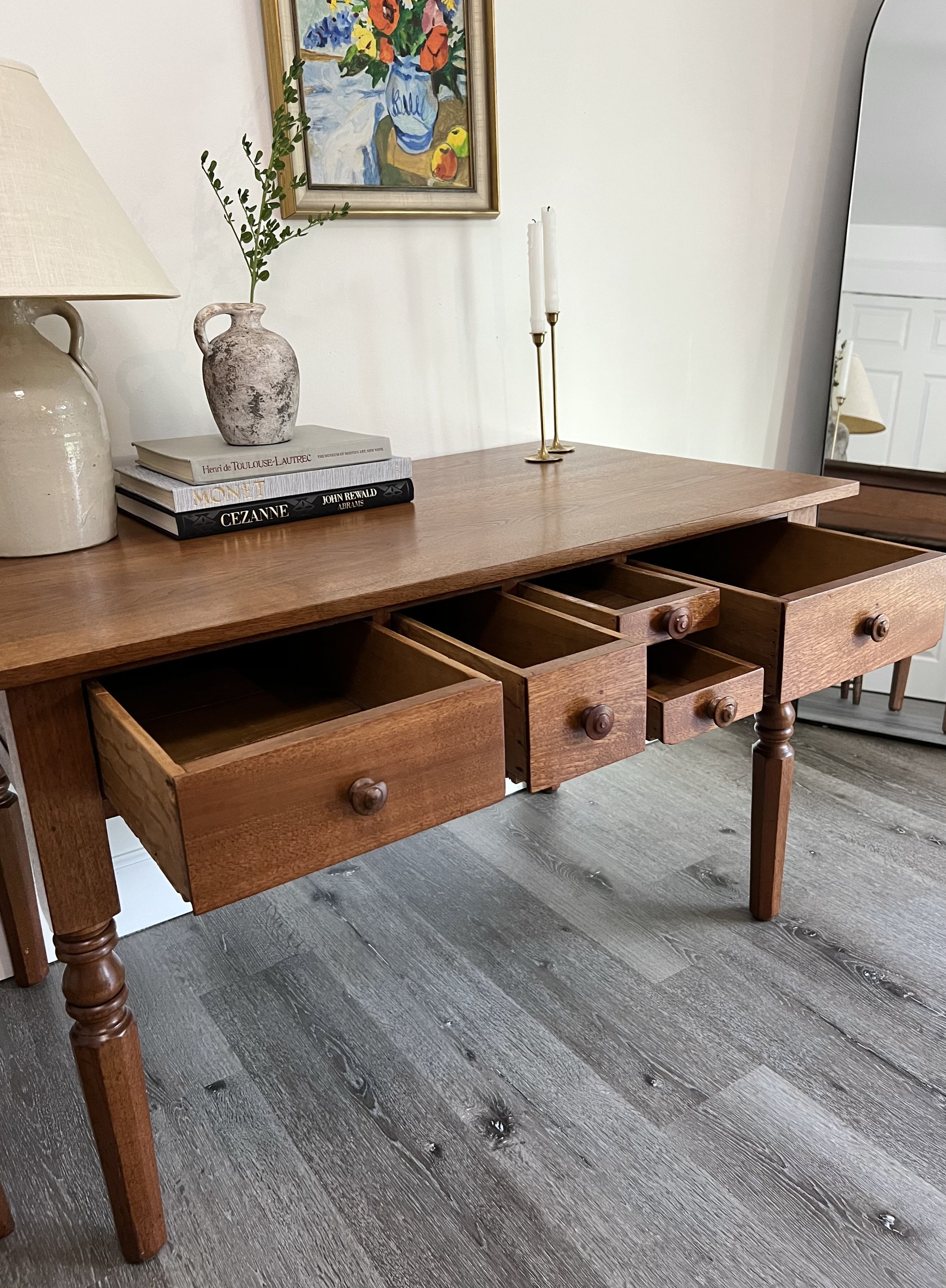 A wooden side table with three open drawers on a grayish wood floor. On top of the table are a beige lamp, a small vase with greenery, a stack of books, and two brass candlesticks with white candles. Above the table is a colorful floral still life pa