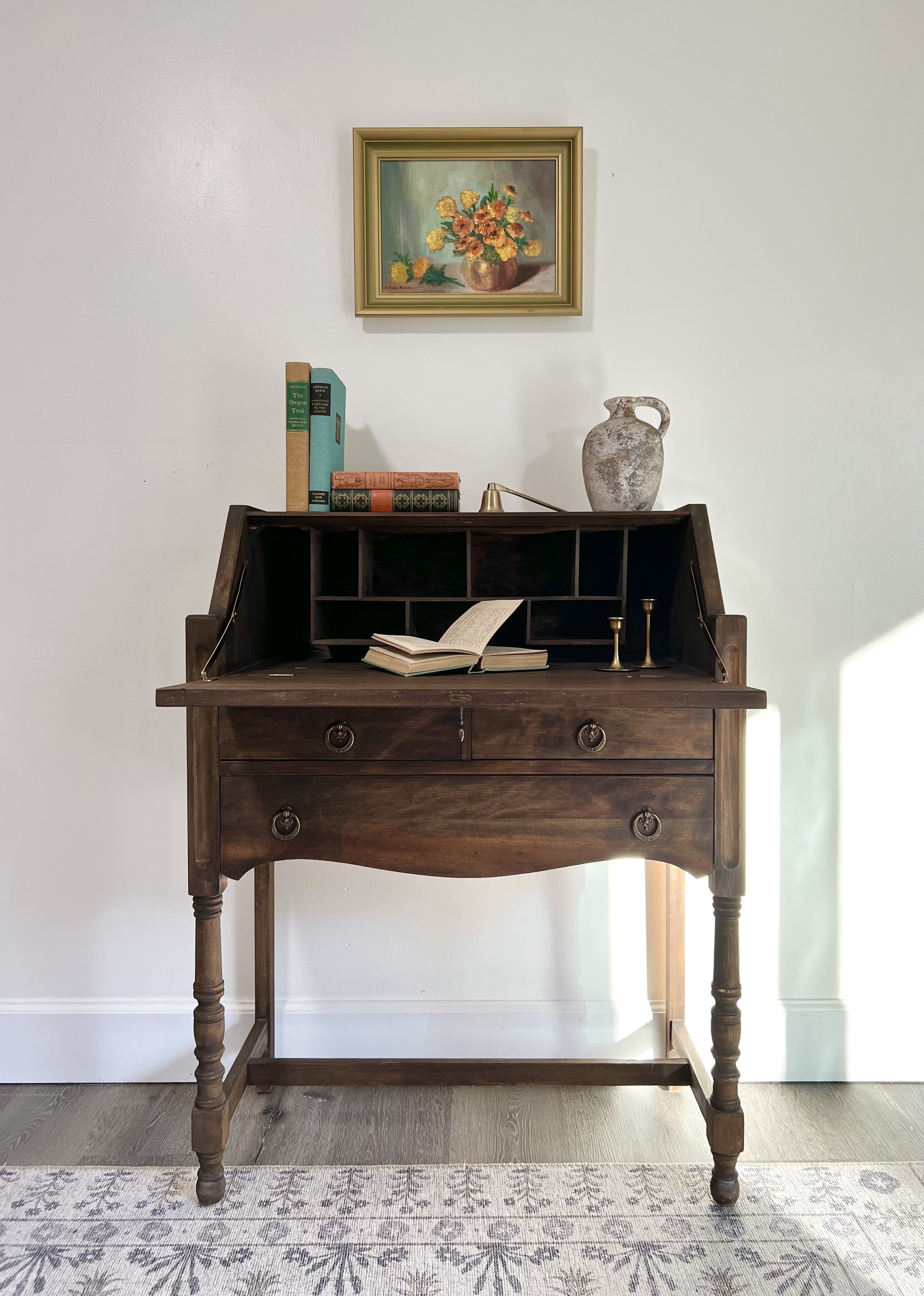 A wooden writing desk with open books, a small brass candlestick, and decorative objects, against a white wall with a framed still life painting of flowers hanging above.