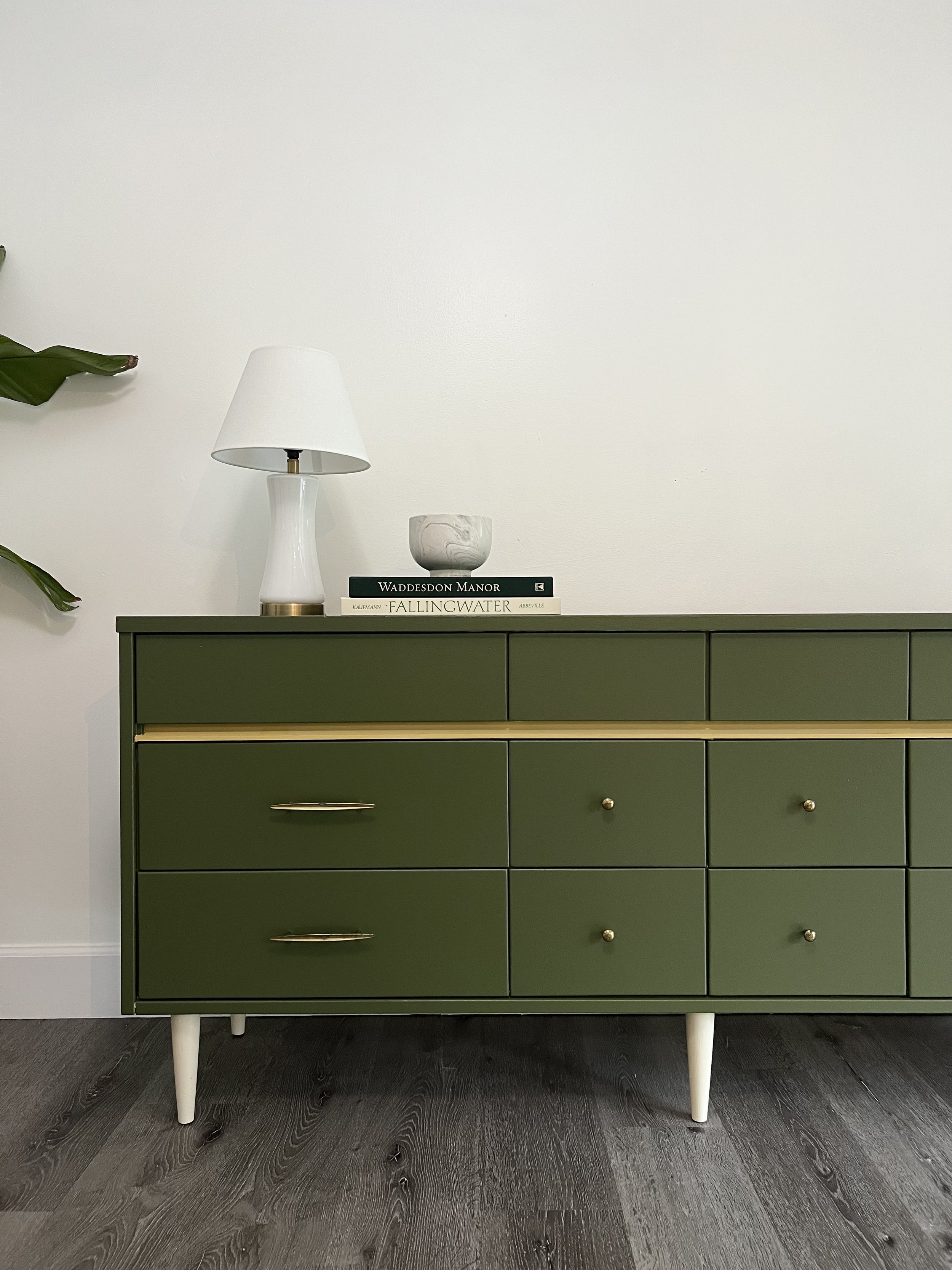 A green mid-century modern dresser with six drawers and white tapered legs, topped with a white table lamp, a marble bowl, and three books, against a plain white wall with dark wood flooring.