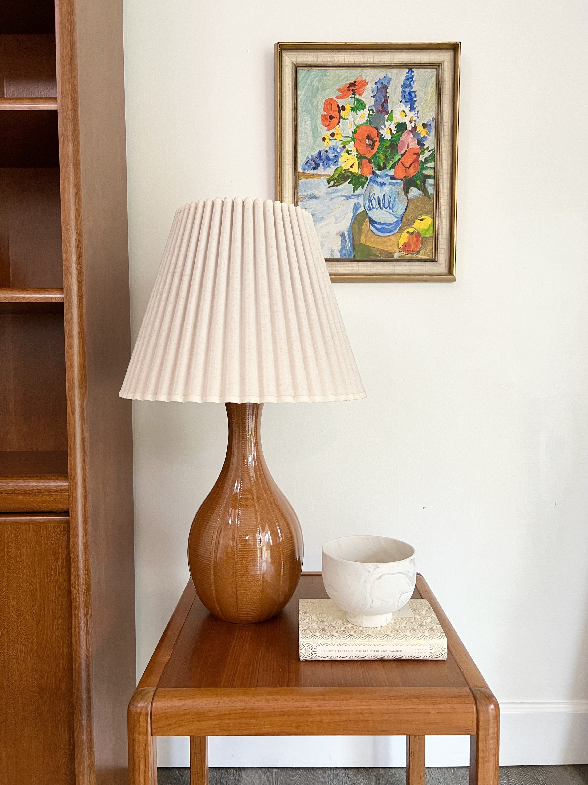 A wooden side table with a large brown ceramic lamp, a white marble bowl, and a closed book. Behind the table, there is a framed colorful floral painting on a white wall.