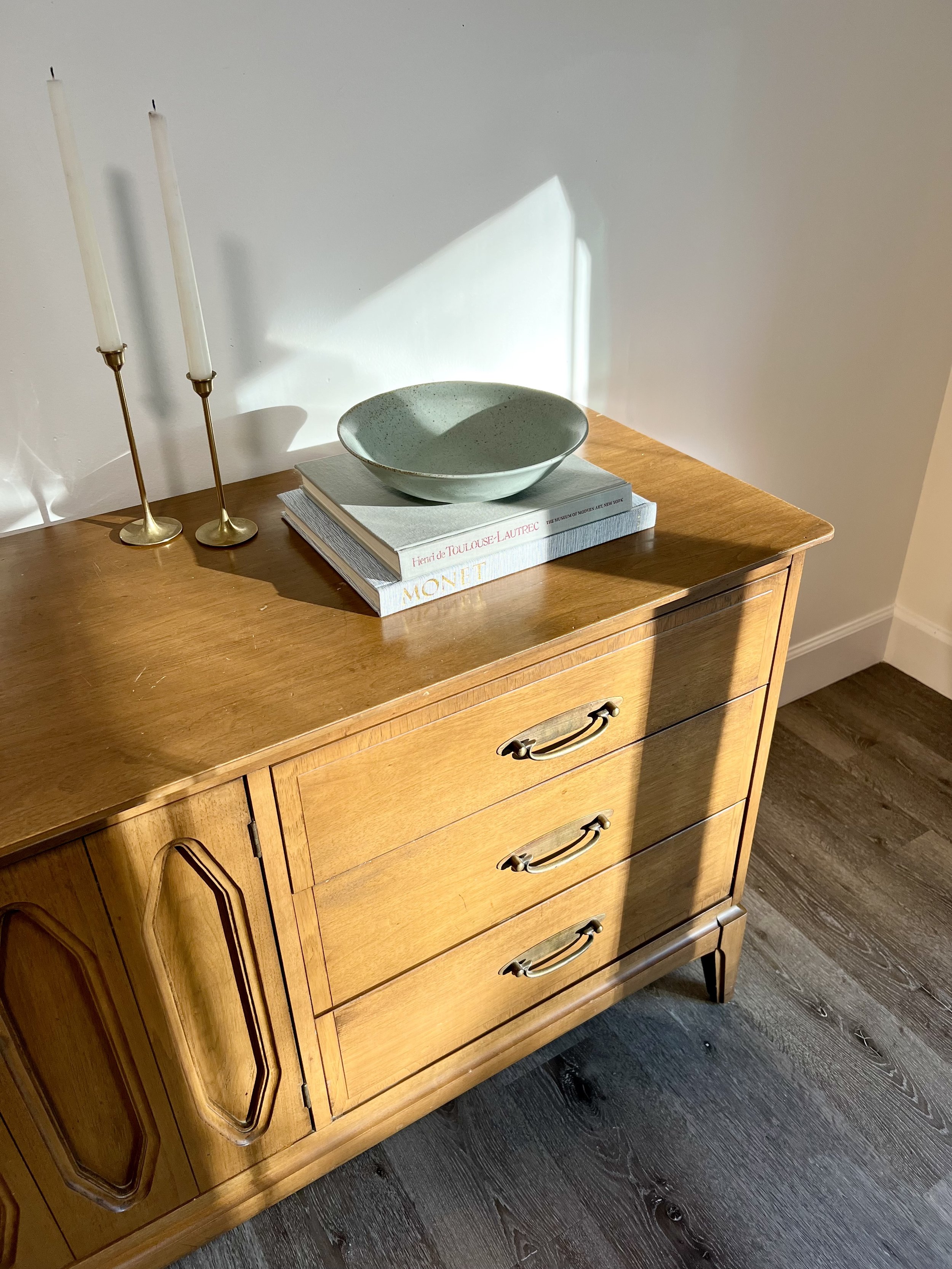 Wooden sideboard with three drawers, two brass handle pulls, and decorative corner details, topped with two brass candlesticks with white candles, a green ceramic bowl, and two stacked books, in a room with wood flooring.