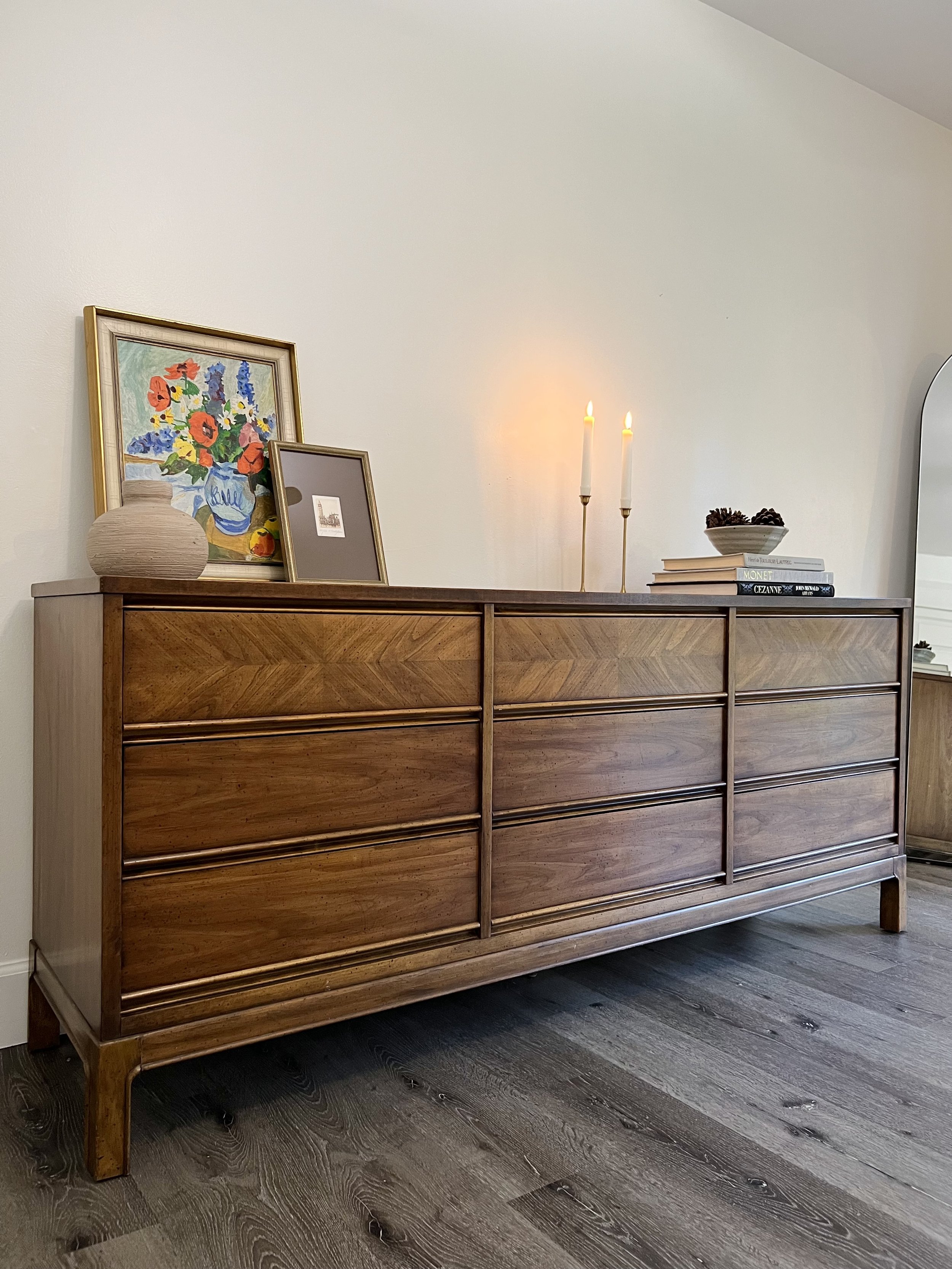 Wooden sideboard with decorative items including framed art, a vase, lit candles, and books in a well-lit room.