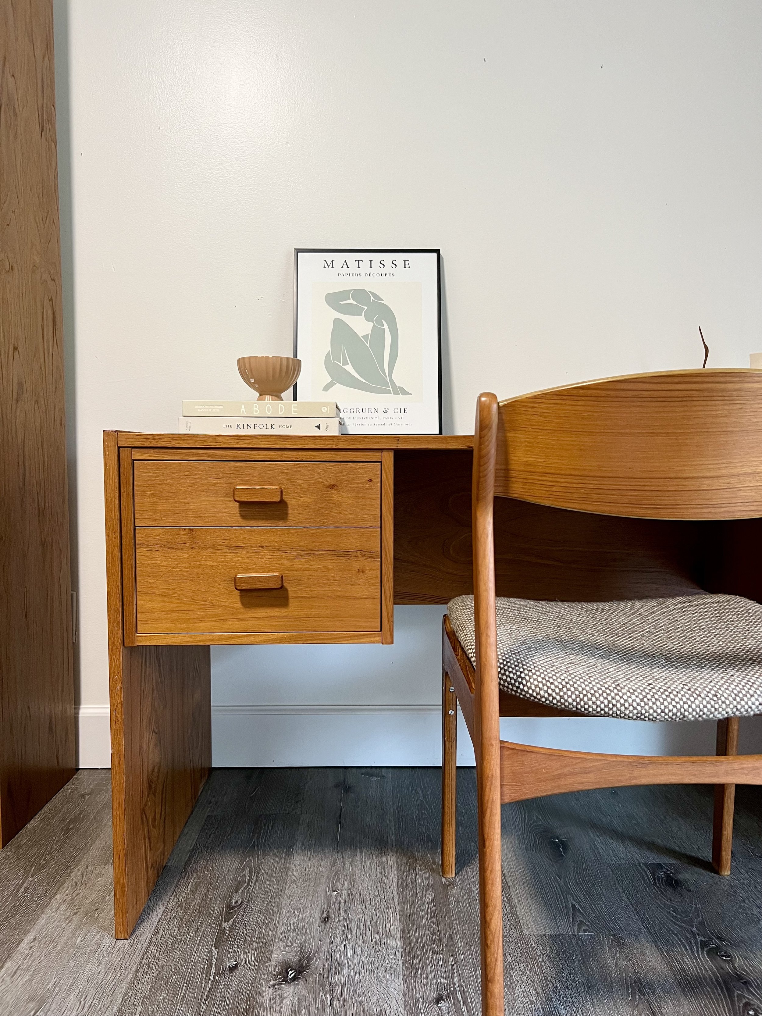 A wooden desk and chair with a framed art print, a bowl, and books on the desk.