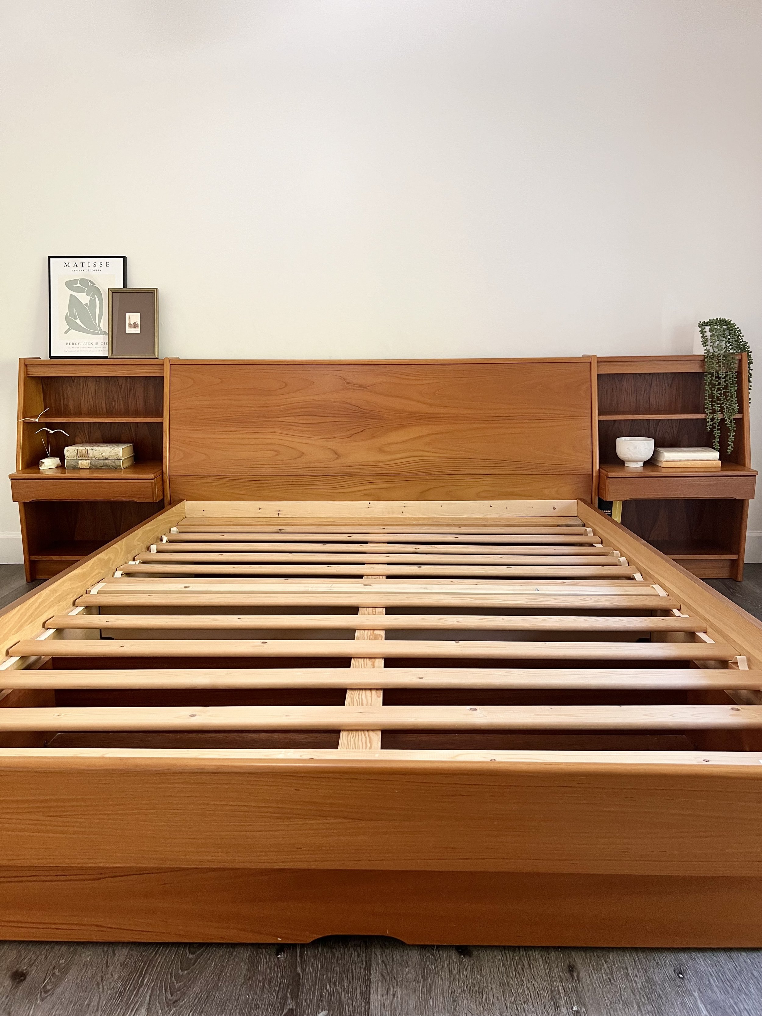 Unmade wooden bed frame with built-in nightstands on either side, decorated with framed art, books, a bowl, and a plant, set against a plain white wall.