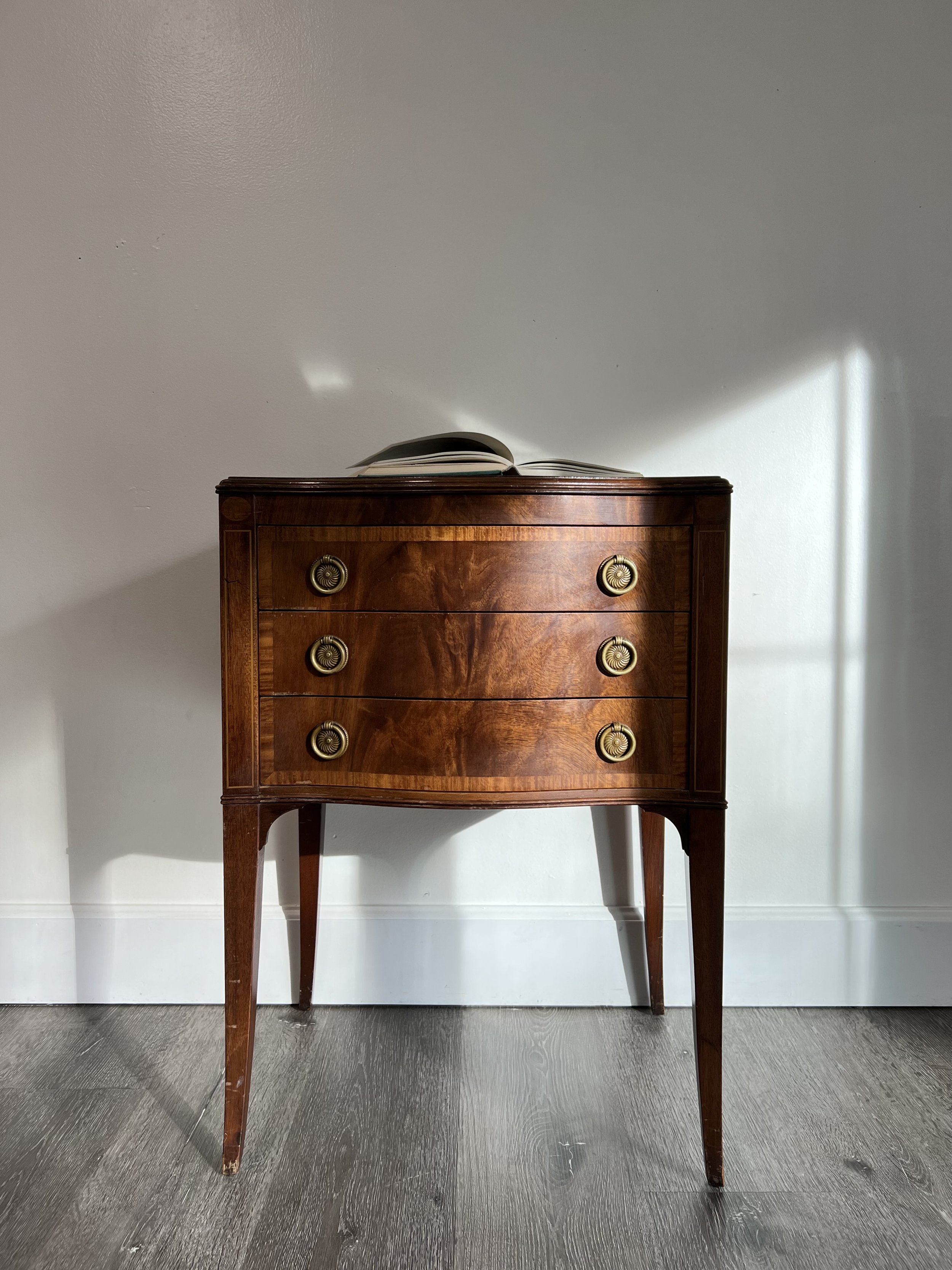 A vintage wooden dresser with four drawers and brass handles, with an open book on top, standing against a white wall with sunlight casting shadows.
