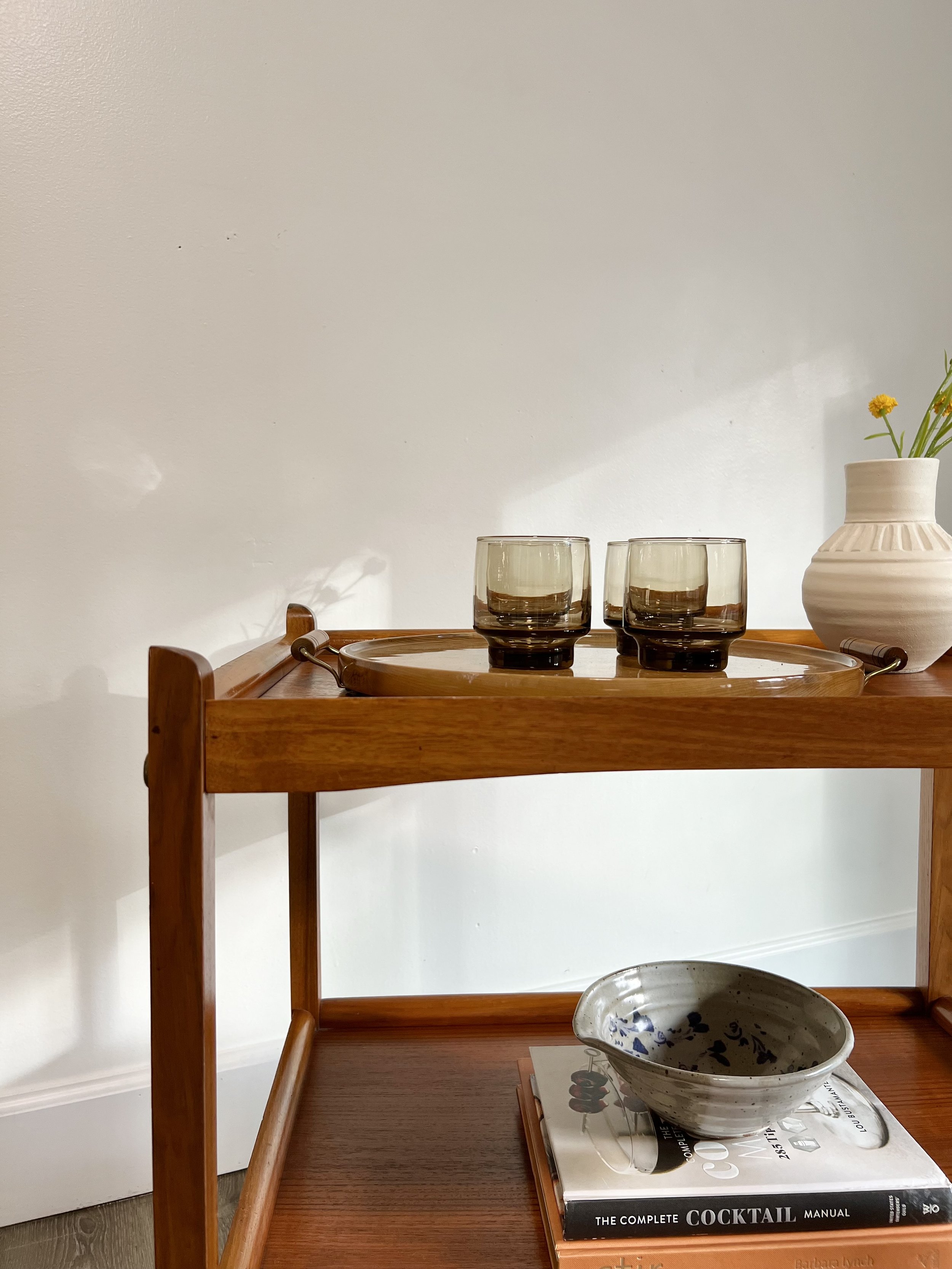 A wooden bar cart with three amber-colored glasses on a round tray, a white vase with yellow flowers, a ceramic bowl, and a cocktail manual book.