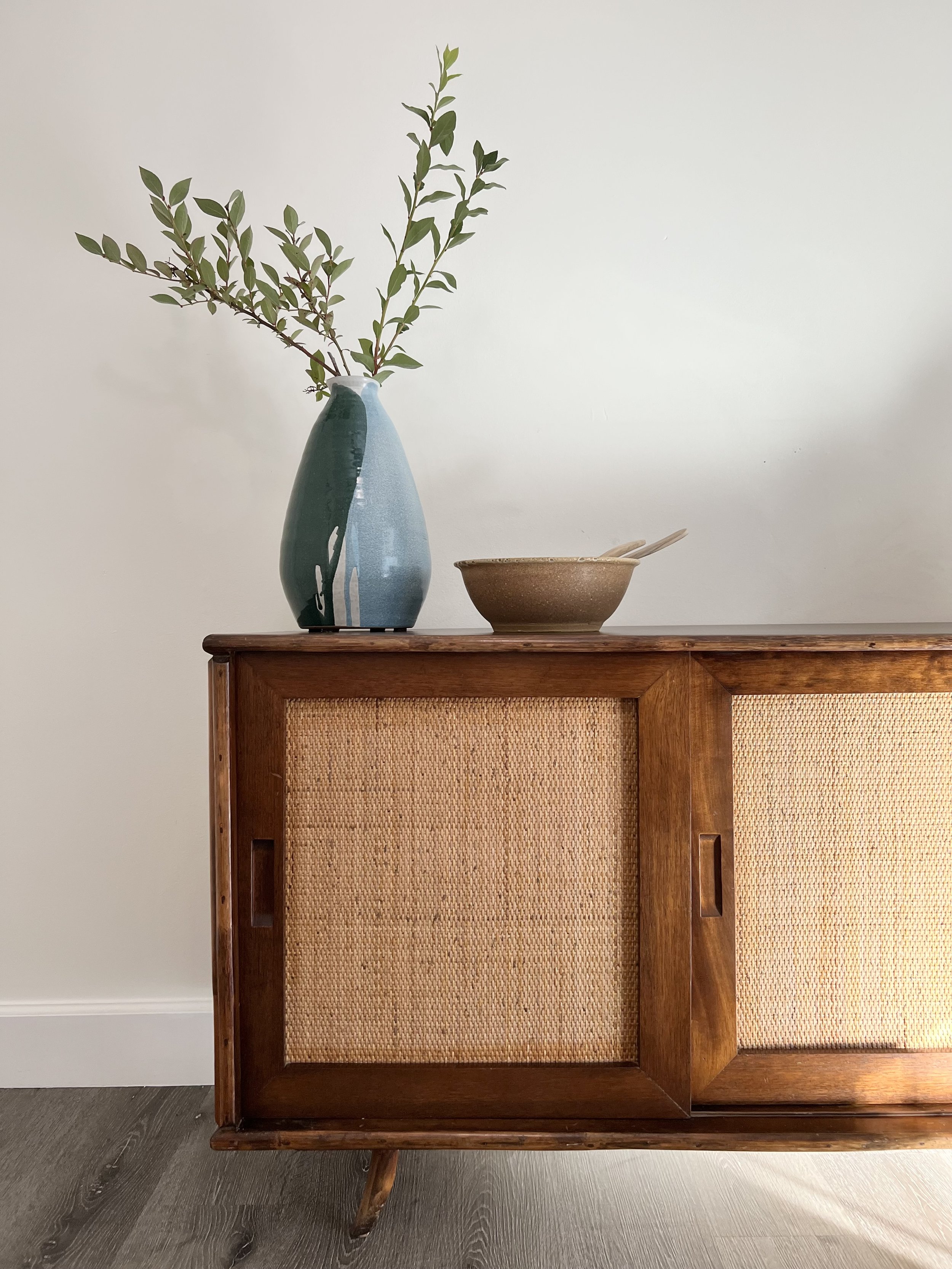A wooden cabinet with woven front panels, a vase with green leafy branches, and a bowl with utensils on top.