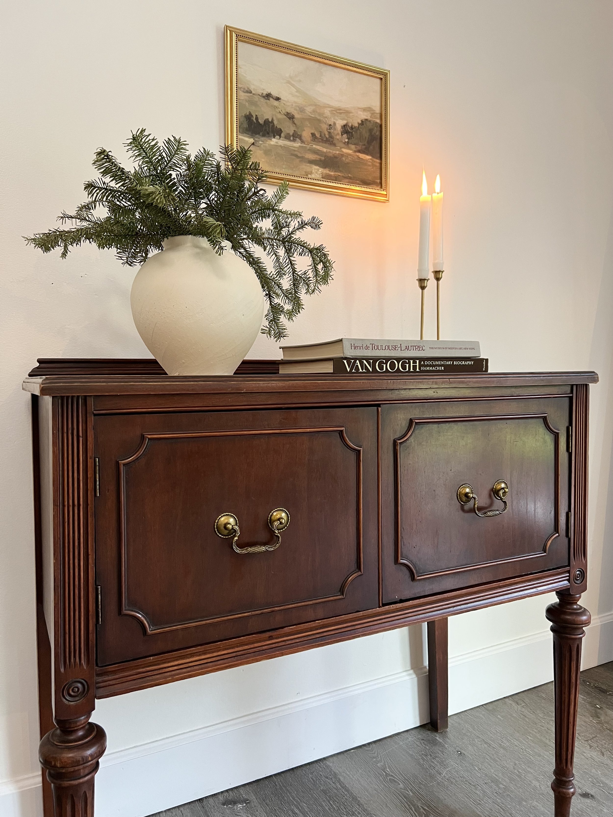 A wooden sideboard with brass handles, topped with a large cream-colored vase filled with pine branches, some books including a Van Gogh biography, and two lit candles in brass holders. A framed landscape painting hangs on the wall above.