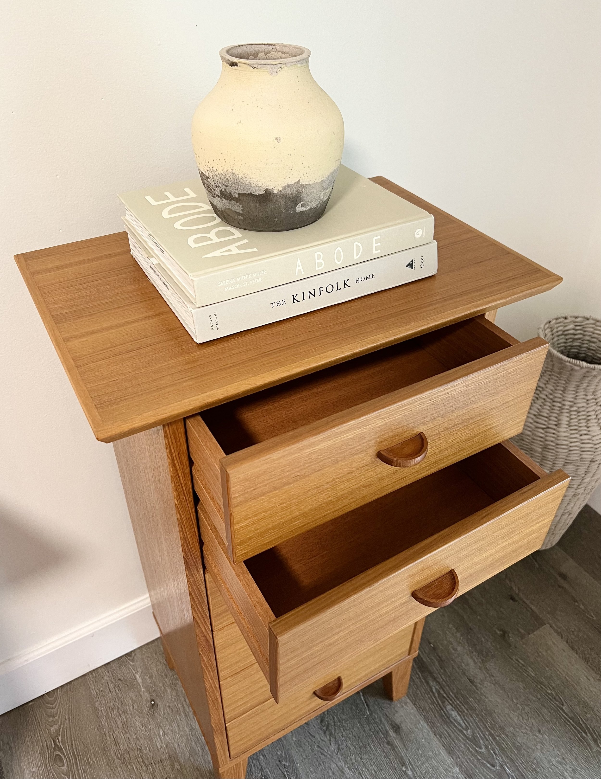 A wooden side table with three drawers, with an open drawer showing interior space. On top of the table are two large hardcover books and a ceramic vase placed on top of the books.