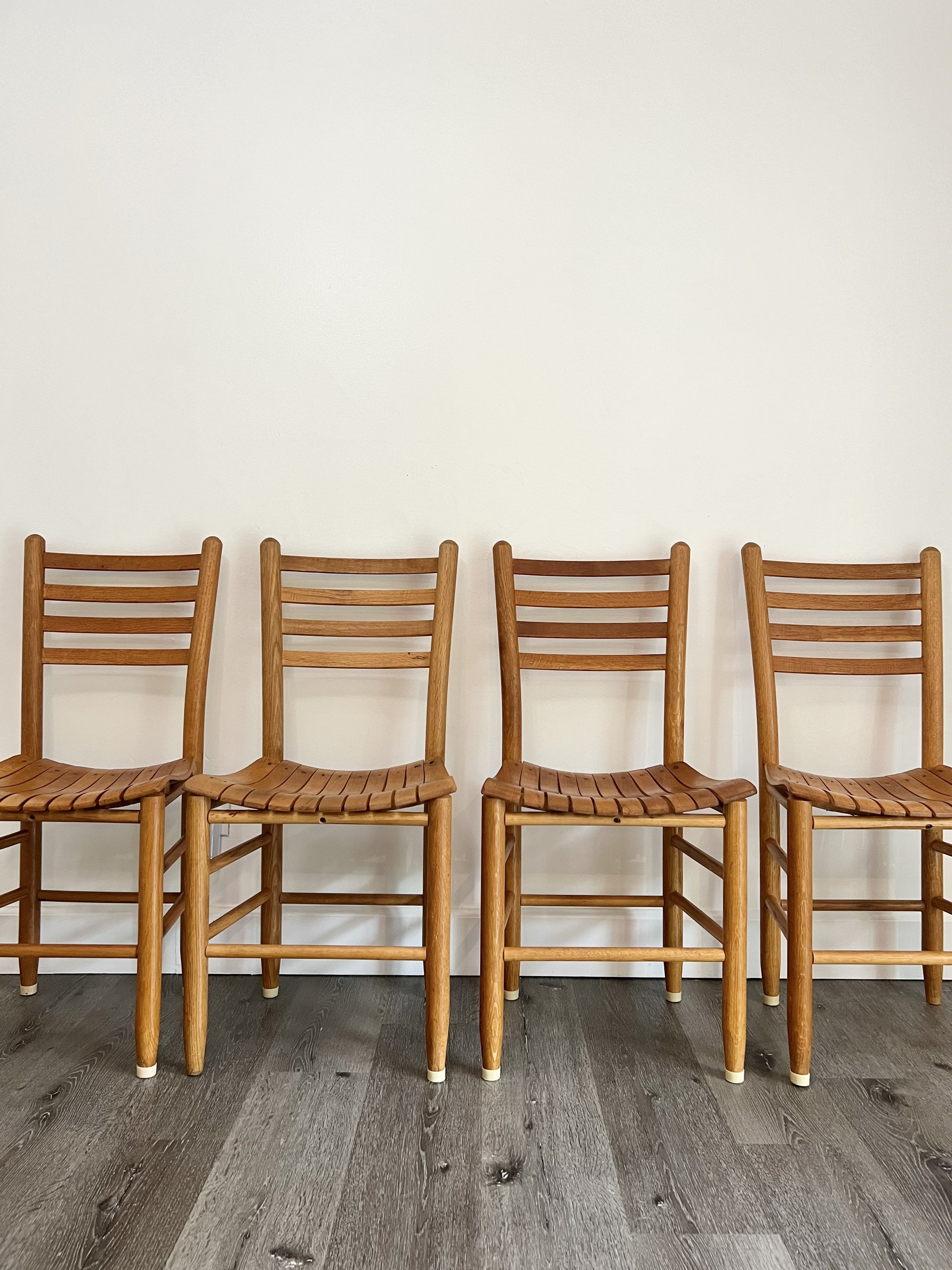 Four wooden chairs with horizontal slats on the backrest and woven wooden seats lined up against a plain white wall, on a dark wooden floor.