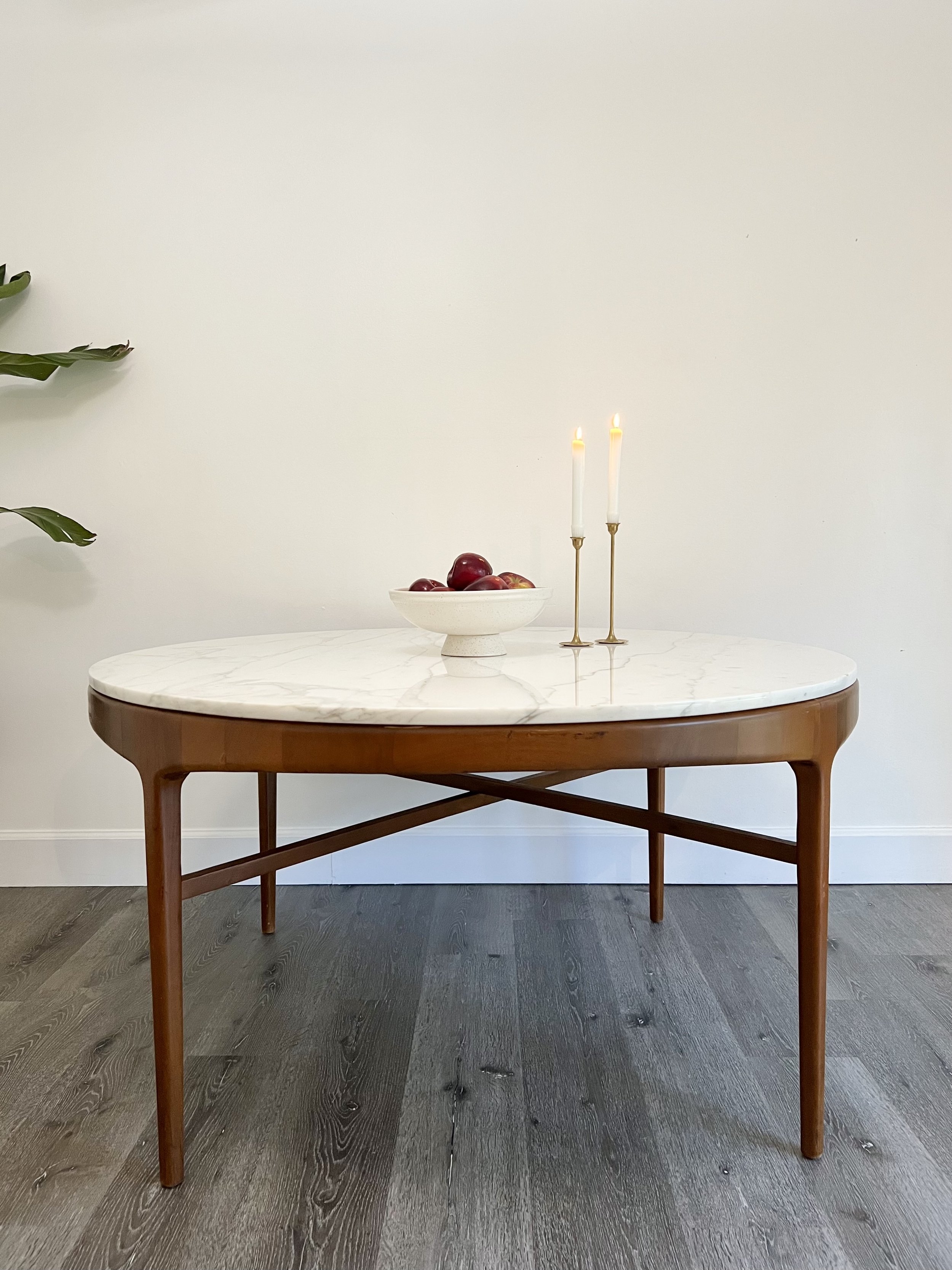 A round white marble-top table with wooden legs, decorated with a bowl of red apples and two lit white candles in gold holders, set against a plain white wall.