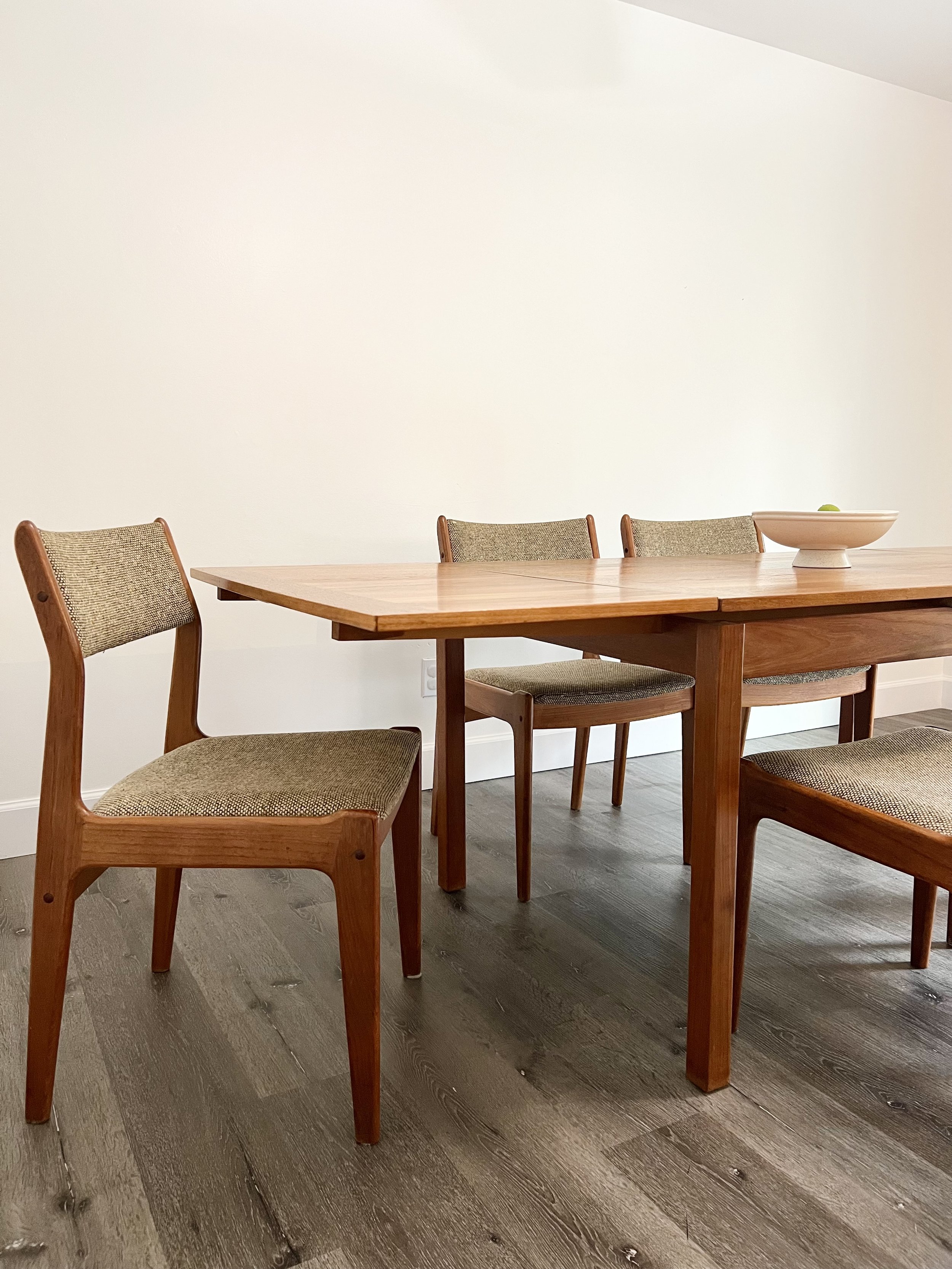 Wood dining table with four upholstered chairs and a white bowl on top, in a minimalist room with white walls and wooden flooring.