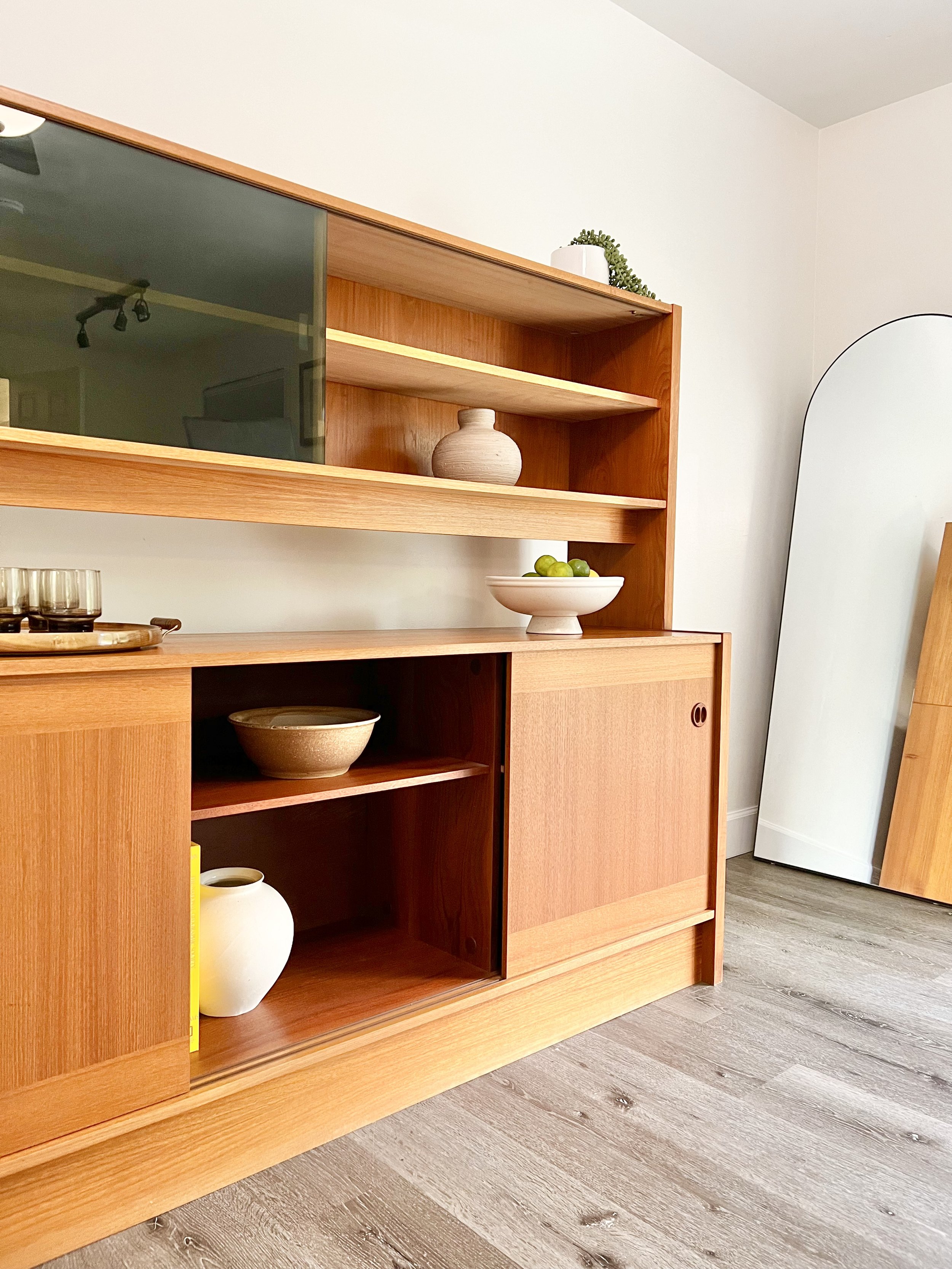 A wooden sideboard with open shelves holding a ceramic bowl, a white vase, and a yellow book. A mirror leaning against the white wall near the corner.