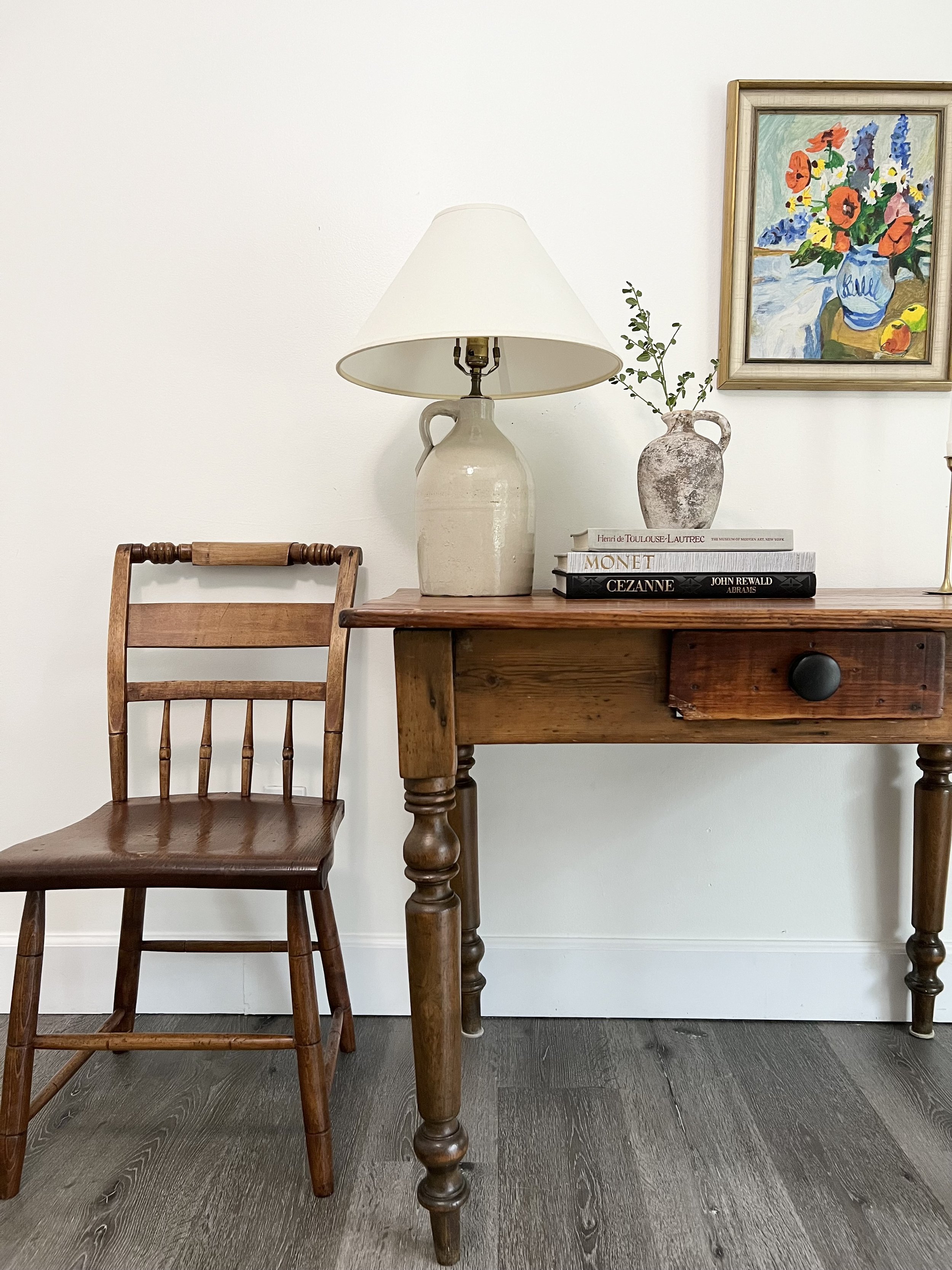A wooden desk with a white ceramic table lamp, a small vase with greenery, and a stack of books next to a framed colorful flower painting on a white wall.