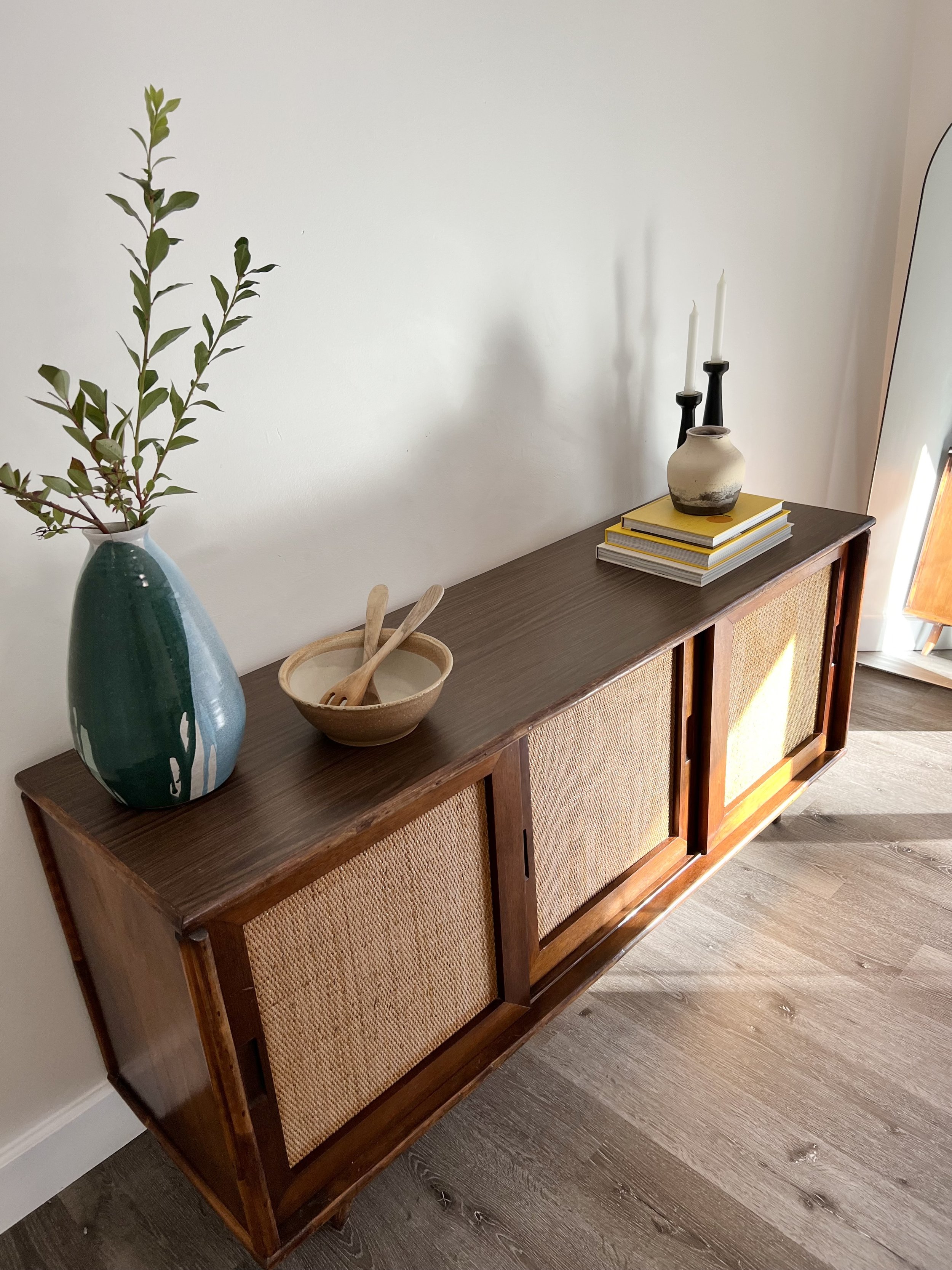 A wooden sideboard with a vase of greenery, a bowl with wooden utensils, and a stack of books with two candlesticks and a decorative vase on top.
