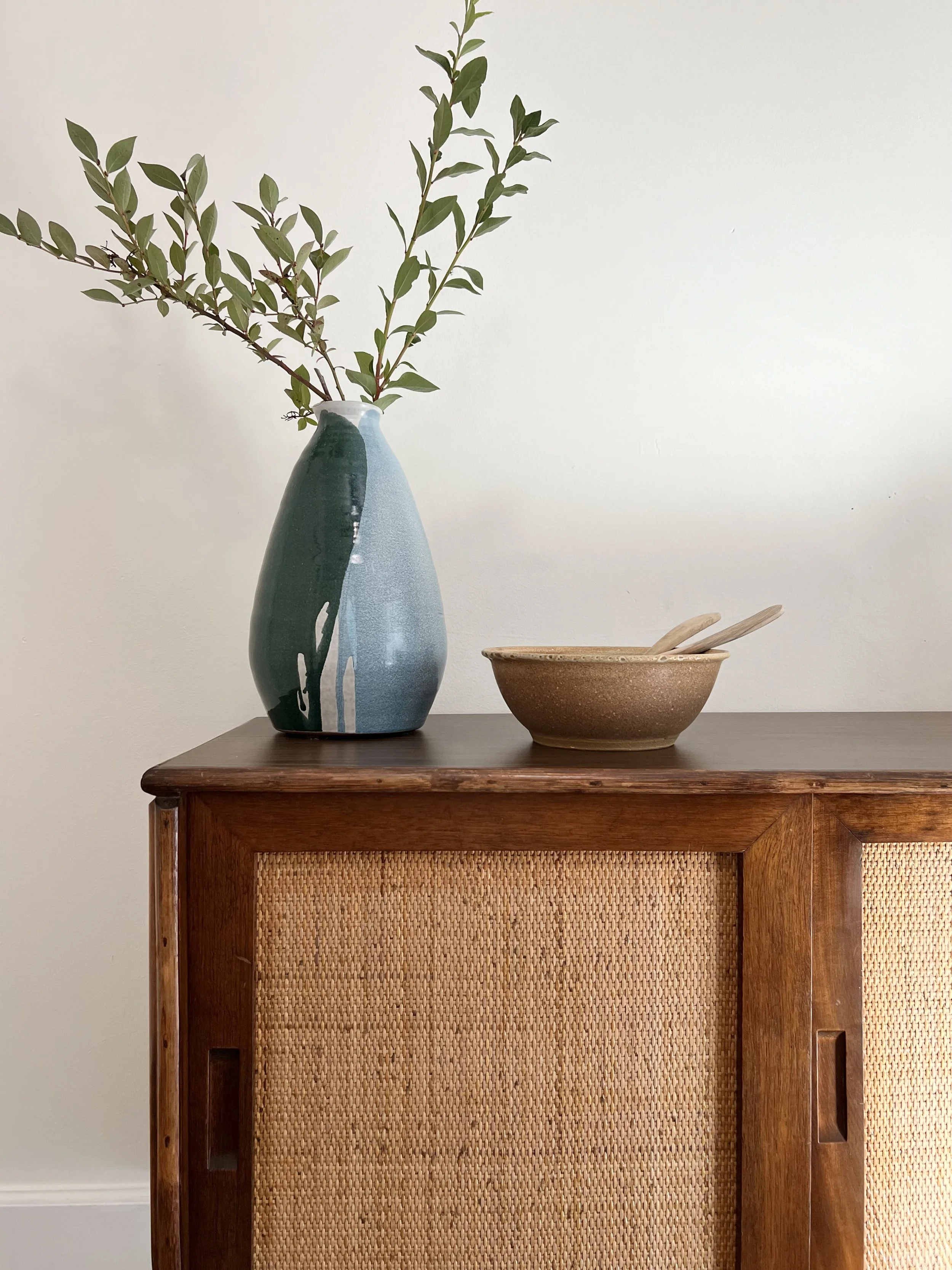 A tall, decorative vase with green and blue abstract design holding green leafy branches, and a small brown bowl with wooden utensils placed on a wooden cabinet with woven details.