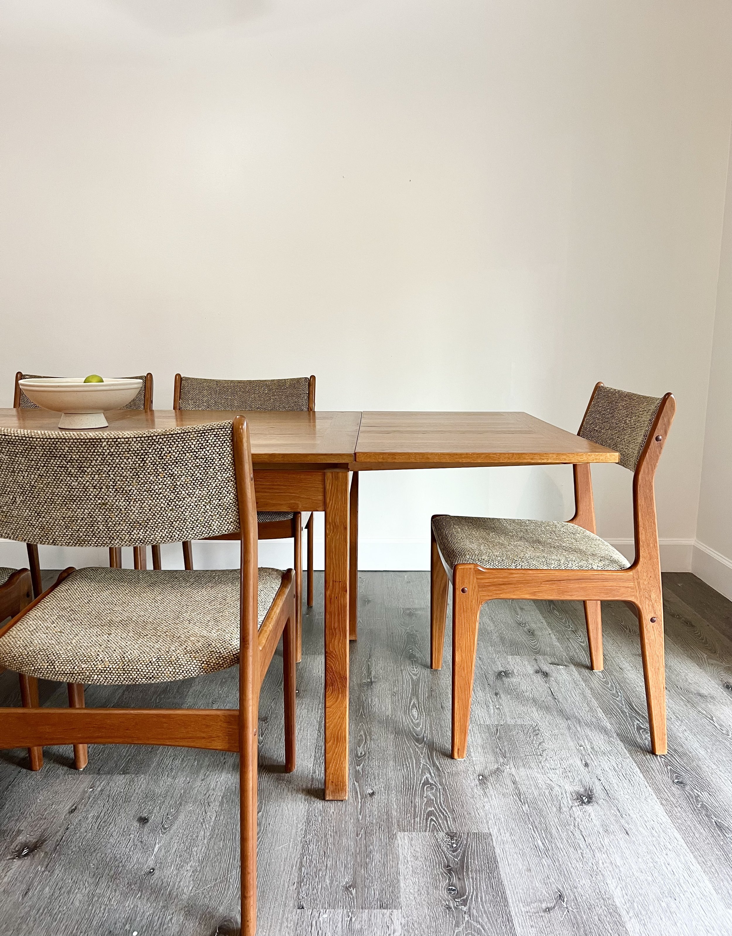 Wooden dining table with four mid-century modern style chairs, one chair is slightly pulled out, and a bowl with green fruit on top, against a plain white wall.