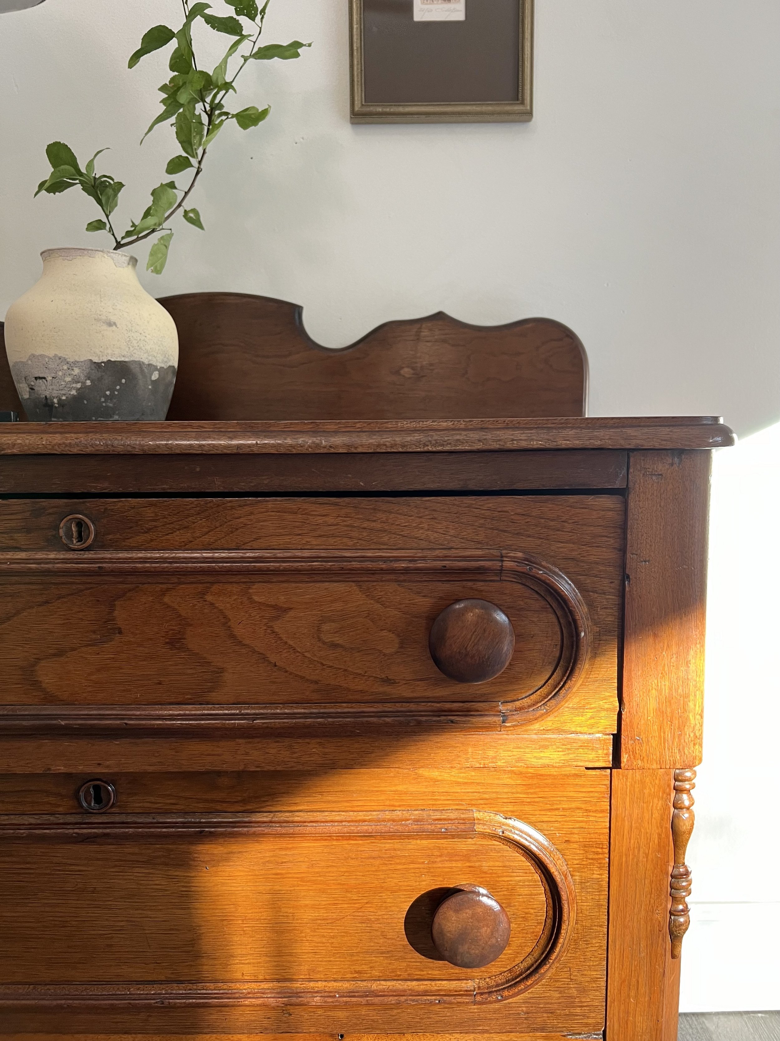 Close-up of a wooden dresser with two drawers, each with round wooden knobs, against a white wall. Sunlight is shining on the dresser. A ceramic vase with a plant is on top of the dresser, and a framed empty corkboard is on the wall above.
