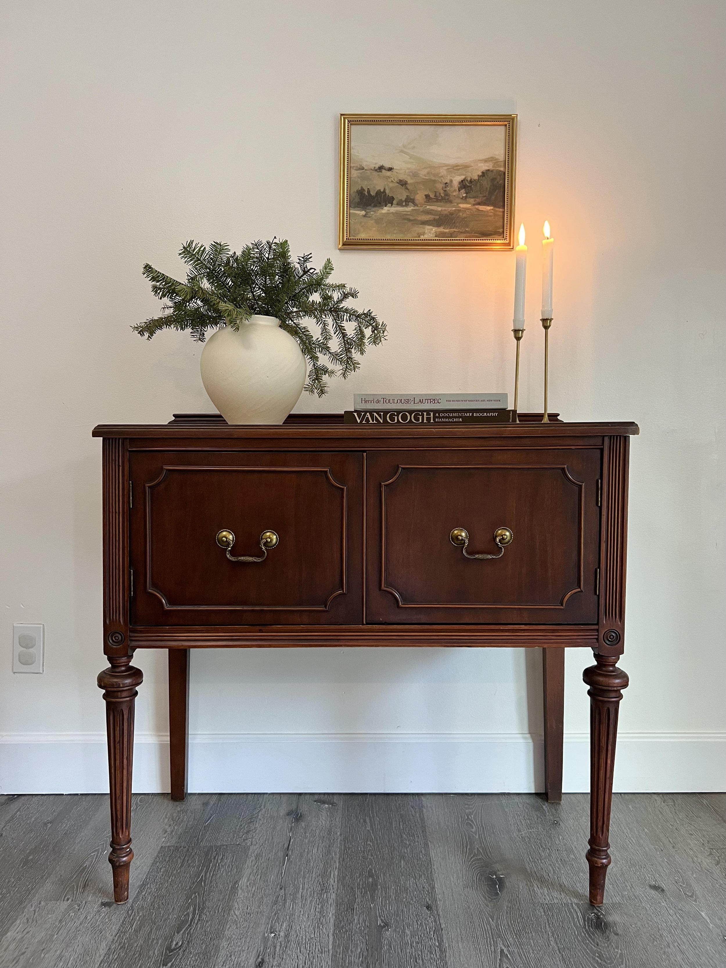 A vintage wooden sideboard with brass handles, decorated with a large ceramic vase filled with pine branches, two lit white candles in brass holders, and a stack of art books, against a white wall with a small landscape painting.
