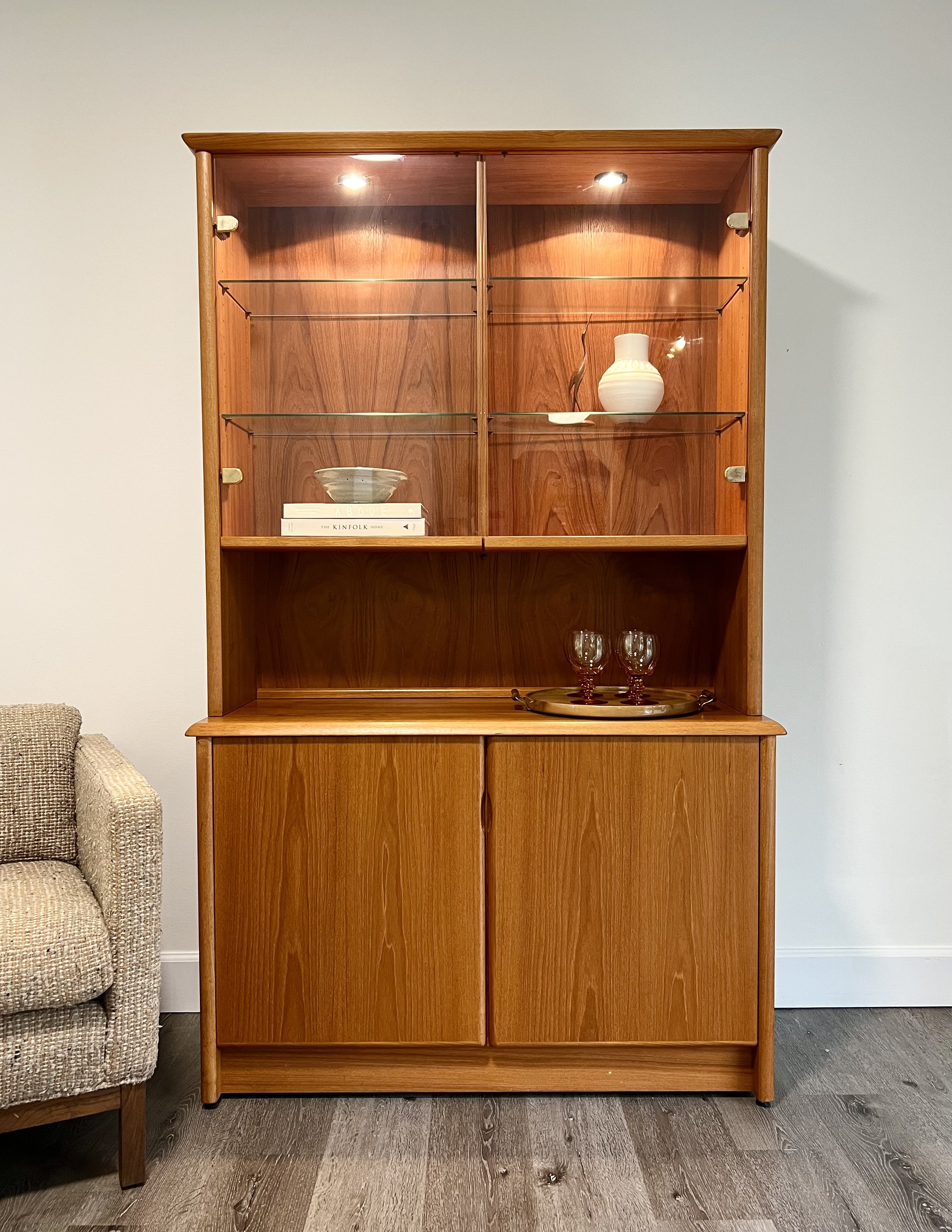 A wooden display cabinet with glass shelves, containing a white vase, a bowl, and books, standing against a plain wall.